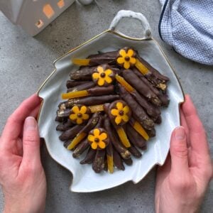 A person holds a leaf-shaped plate filled with chocolate-dipped candied orange peels, decorated with small yellow flower-shaped chocolates.