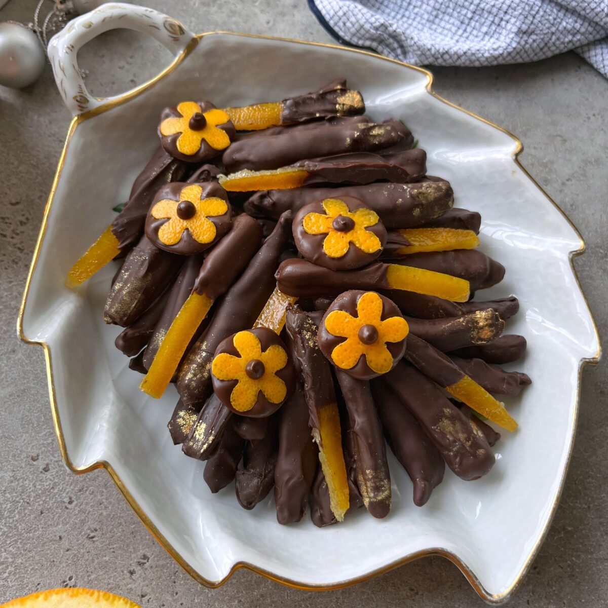 A white leaf-shaped platter holds chocolate-covered orange peels, some topped with small orange flower decorations and gold accents.