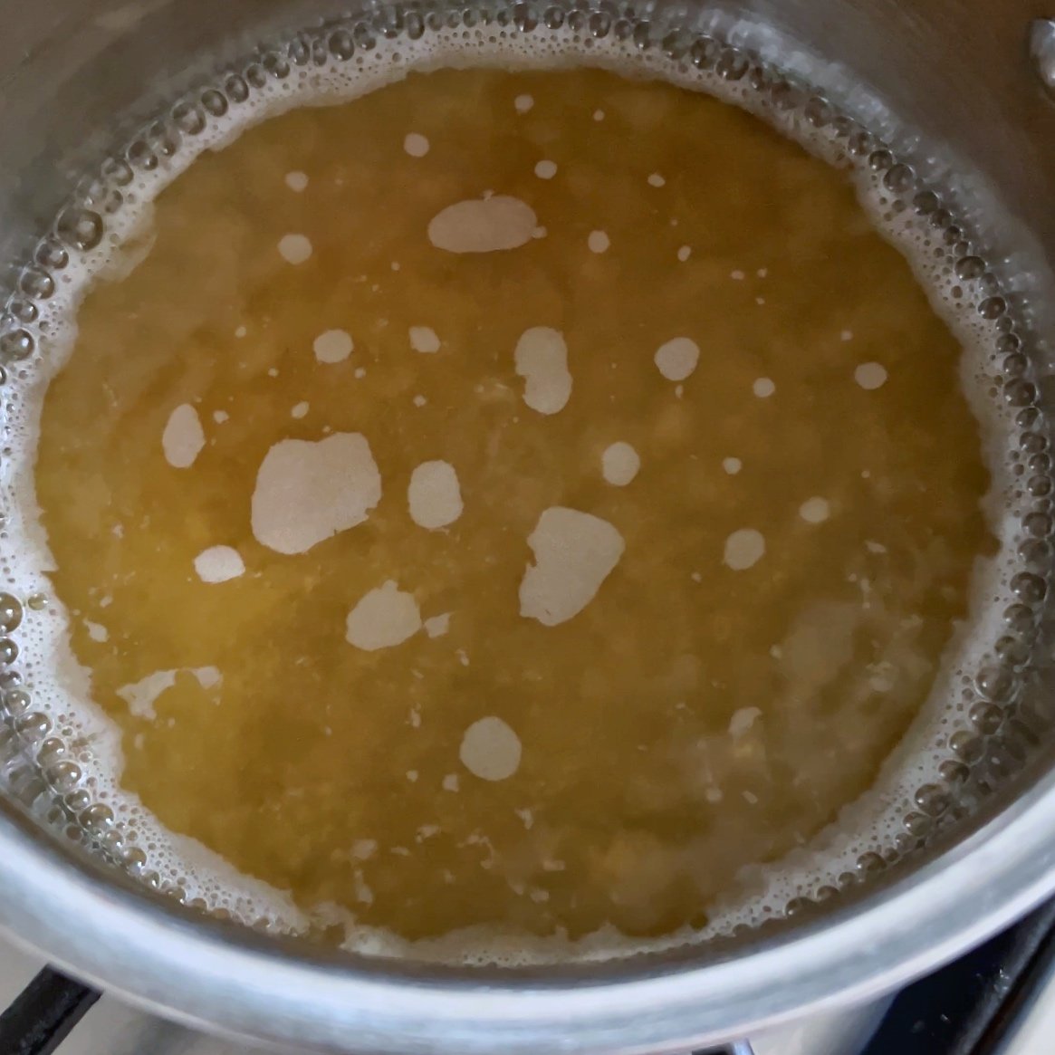 A saucepan containing simple syrup is boiling with foam and bubbles on the surface, heating on a stovetop.