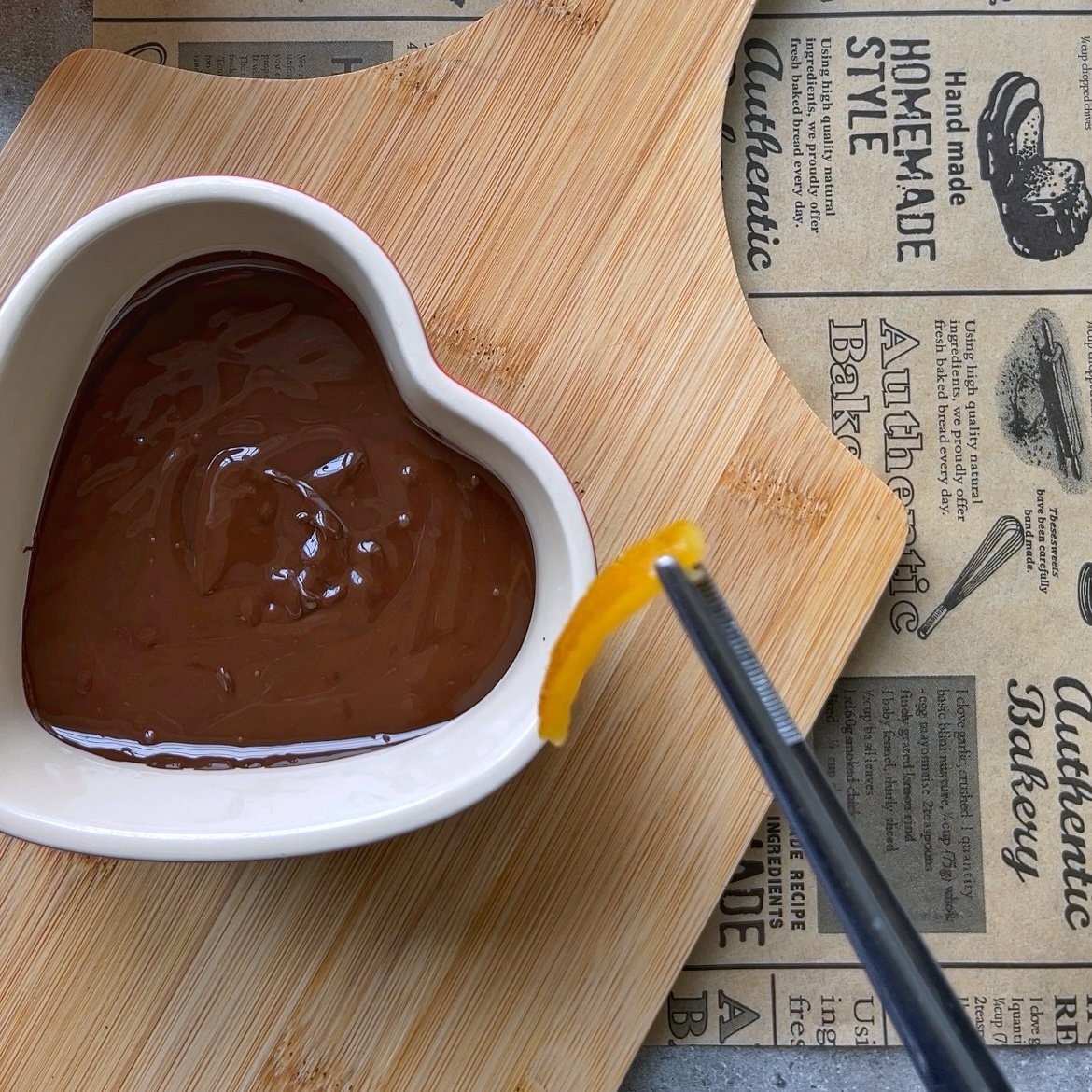A heart-shaped bowl filled with melted chocolate sits on a wooden board, while metal tongs hold a strip of orange peel above it.