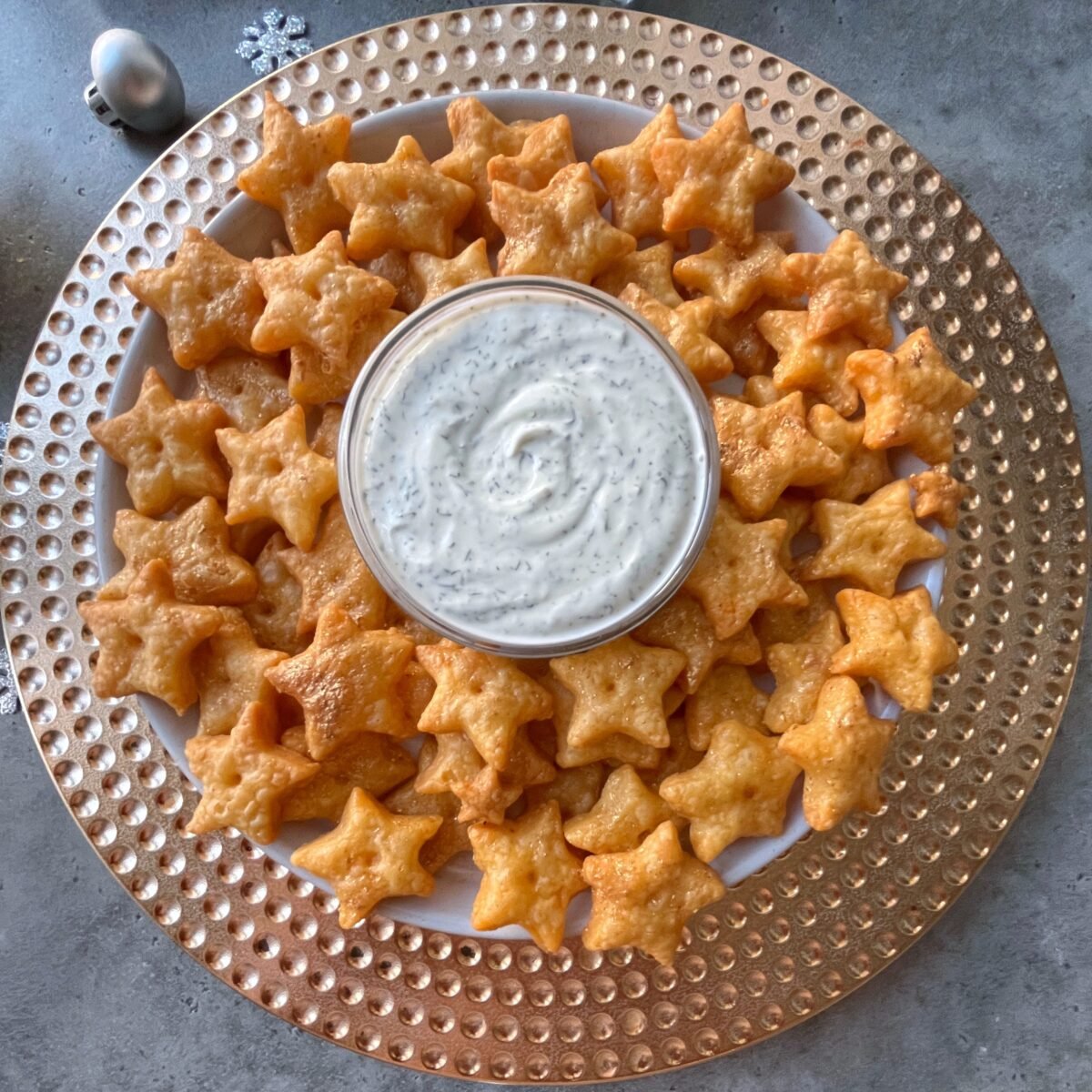 A plate of star-shaped crackers arranged around a bowl of white dipping sauce on a gold-rimmed charger.