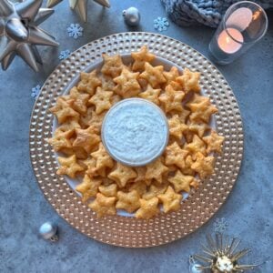 A plate of star-shaped crackers arranged around a bowl of white dip on a gold charger, surrounded by festive holiday decorations and a lit candle.