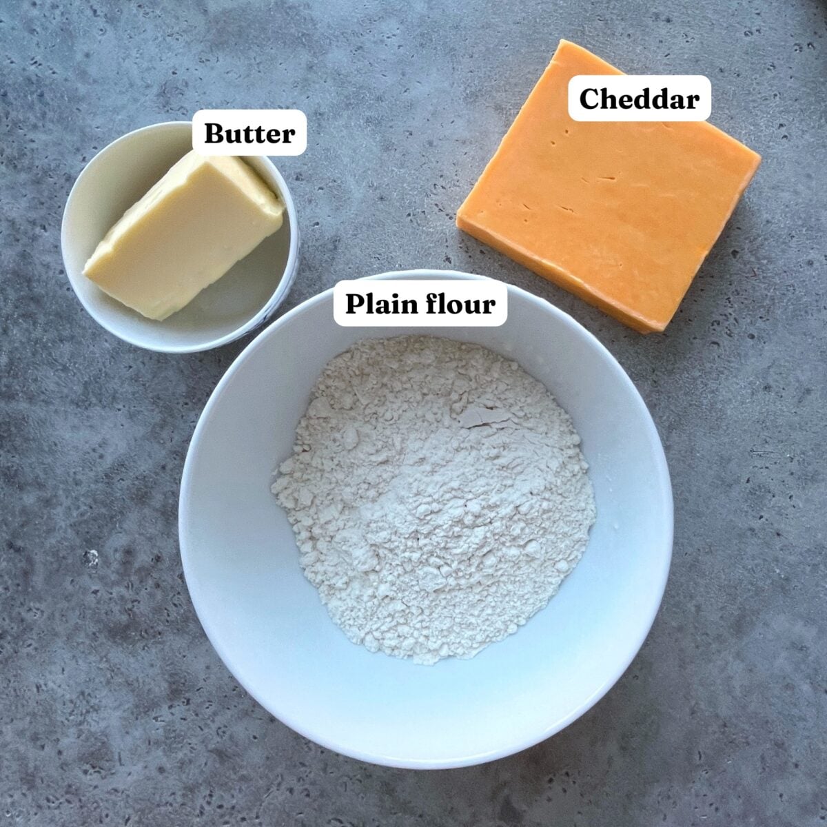 A bowl of plain flour, a block of cheddar cheese, and a dish of butter are arranged on a gray surface.