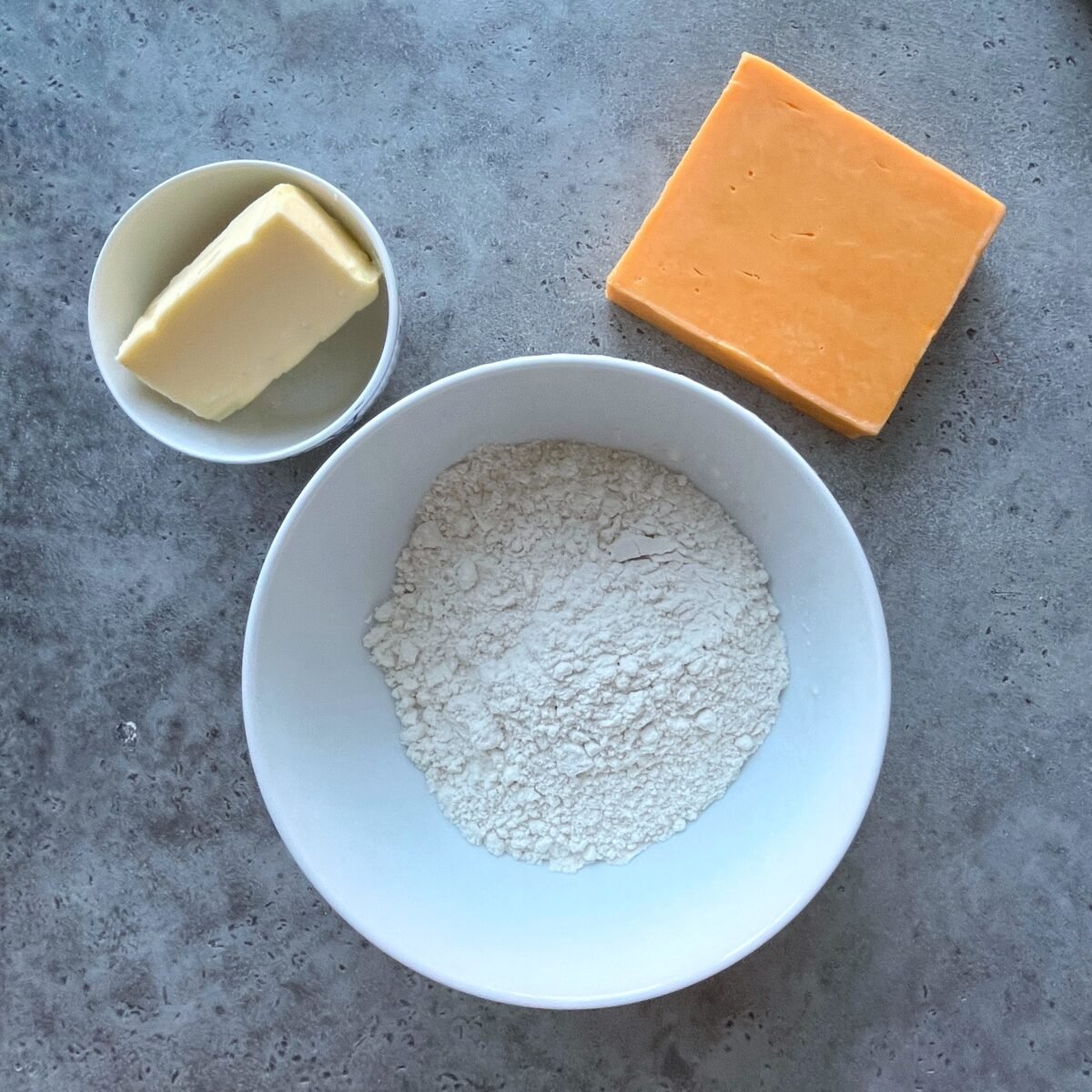 A bowl of flour, a block of cheddar cheese, and a dish of butter on a gray countertop.