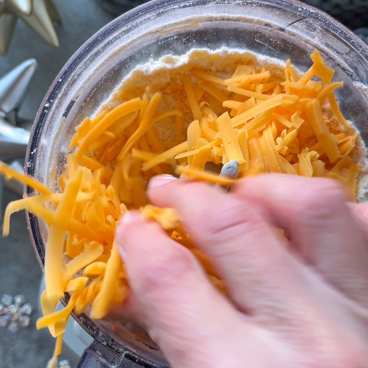 A hand places shredded cheddar cheese into a food processor containing flour, preparing the dough for homemade Cheddar Crackers.