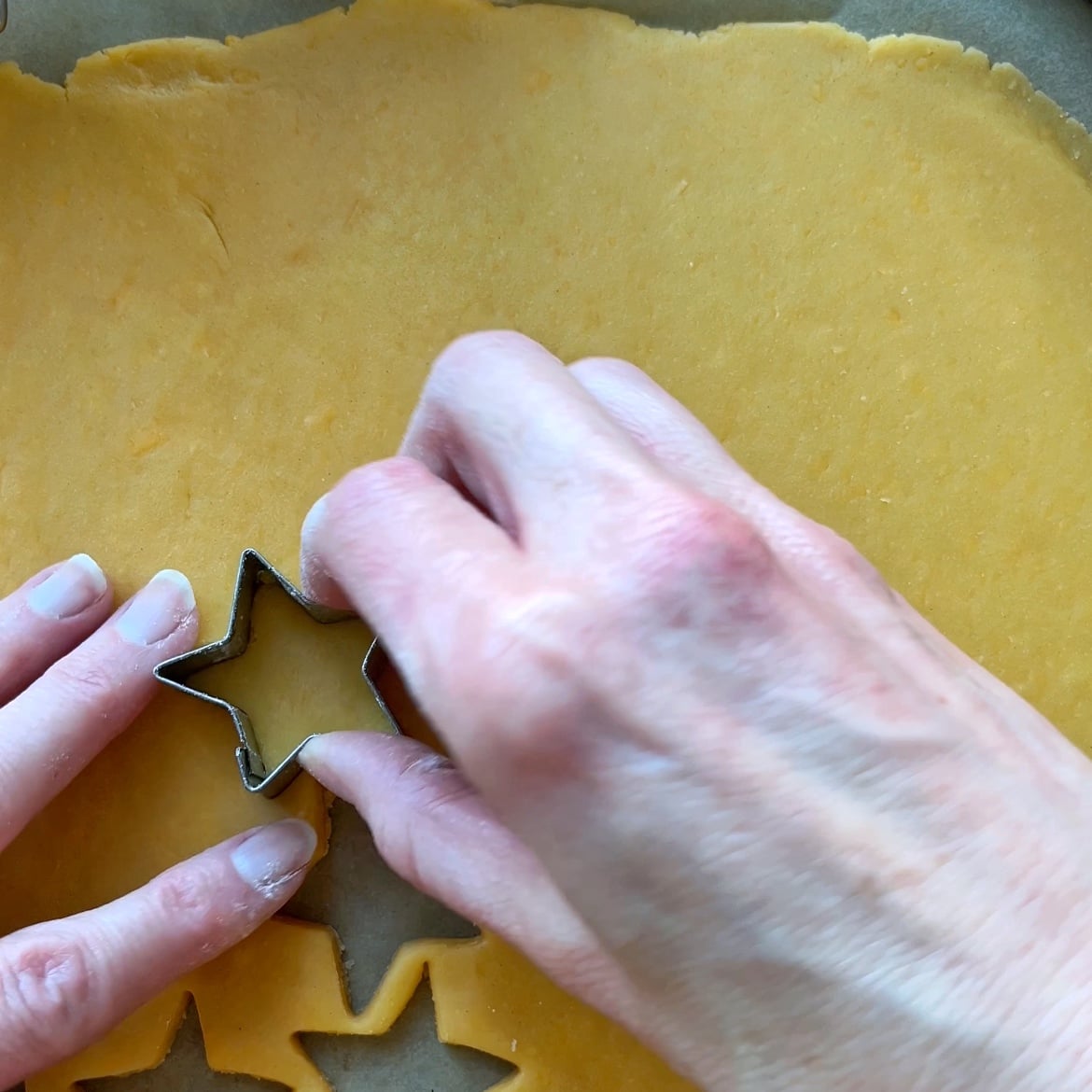 Hands using a metal star-shaped cookie cutter to cut shapes from rolled-out cheddar cracker dough on a flat surface.