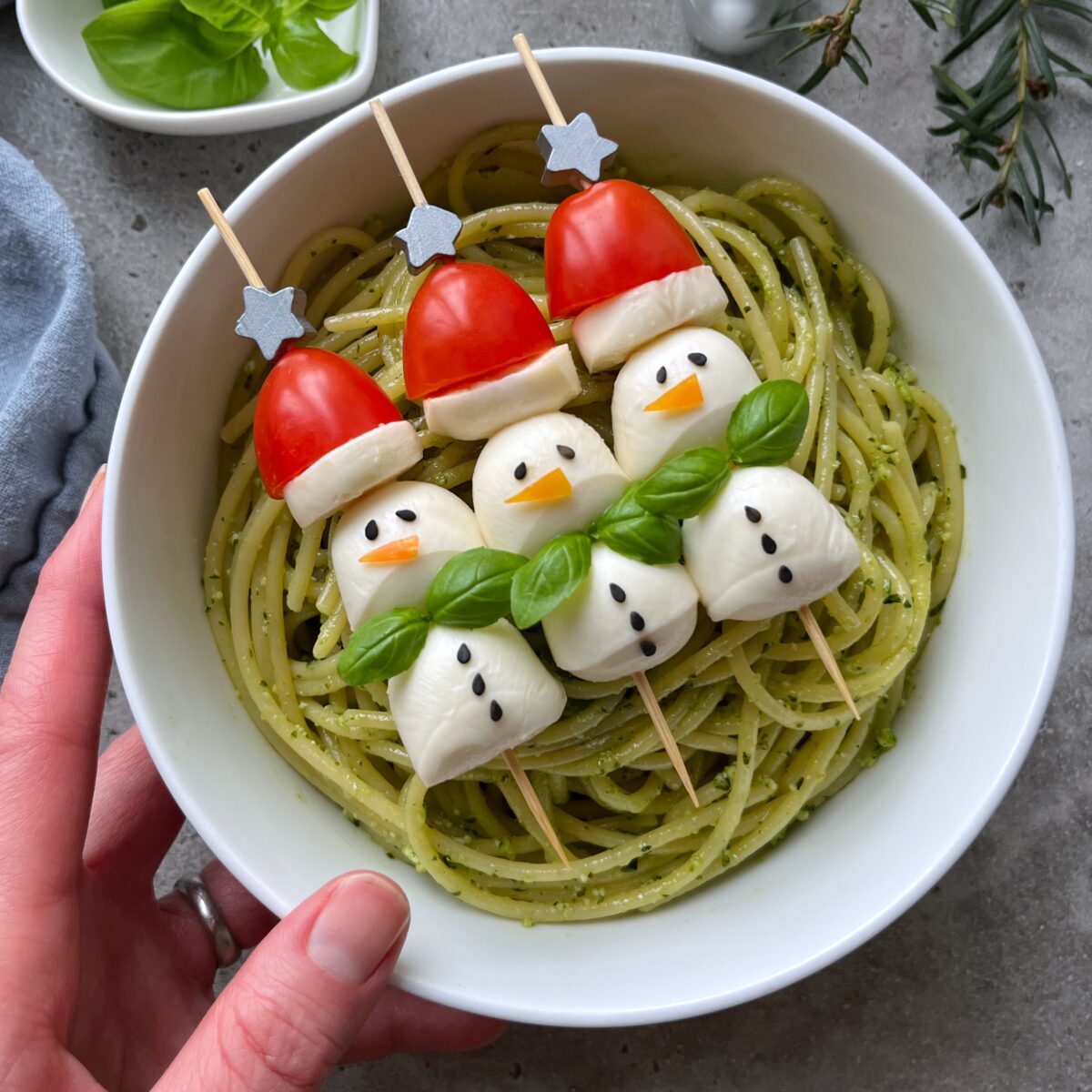 A hand holding a bowl of pasta with pesto topped with skewers shaped like snowmen and Santa hats made from mozzarella, tomatoes, black sesame seeds, and basil.