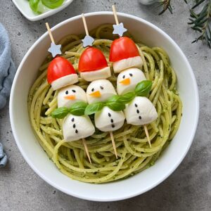 Bowl of pesto pasta topped with skewers of mozzarella and tomato snowmen, decorated with basil leaves and black sesame seeds for facial features and buttons.
