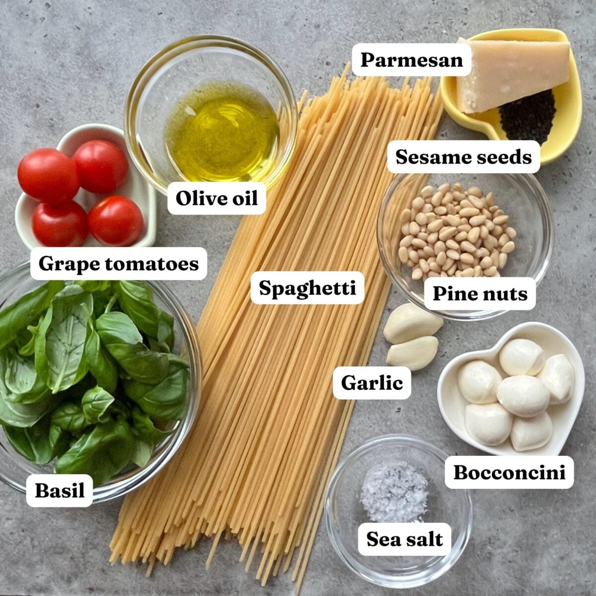 A flat lay of Christmas Pasta ingredients featuring spaghetti, grape tomatoes, basil, Parmesan, olive oil, sesame seeds, pine nuts, garlic, bocconcini, and sea salt, each festive ingredient labeled.