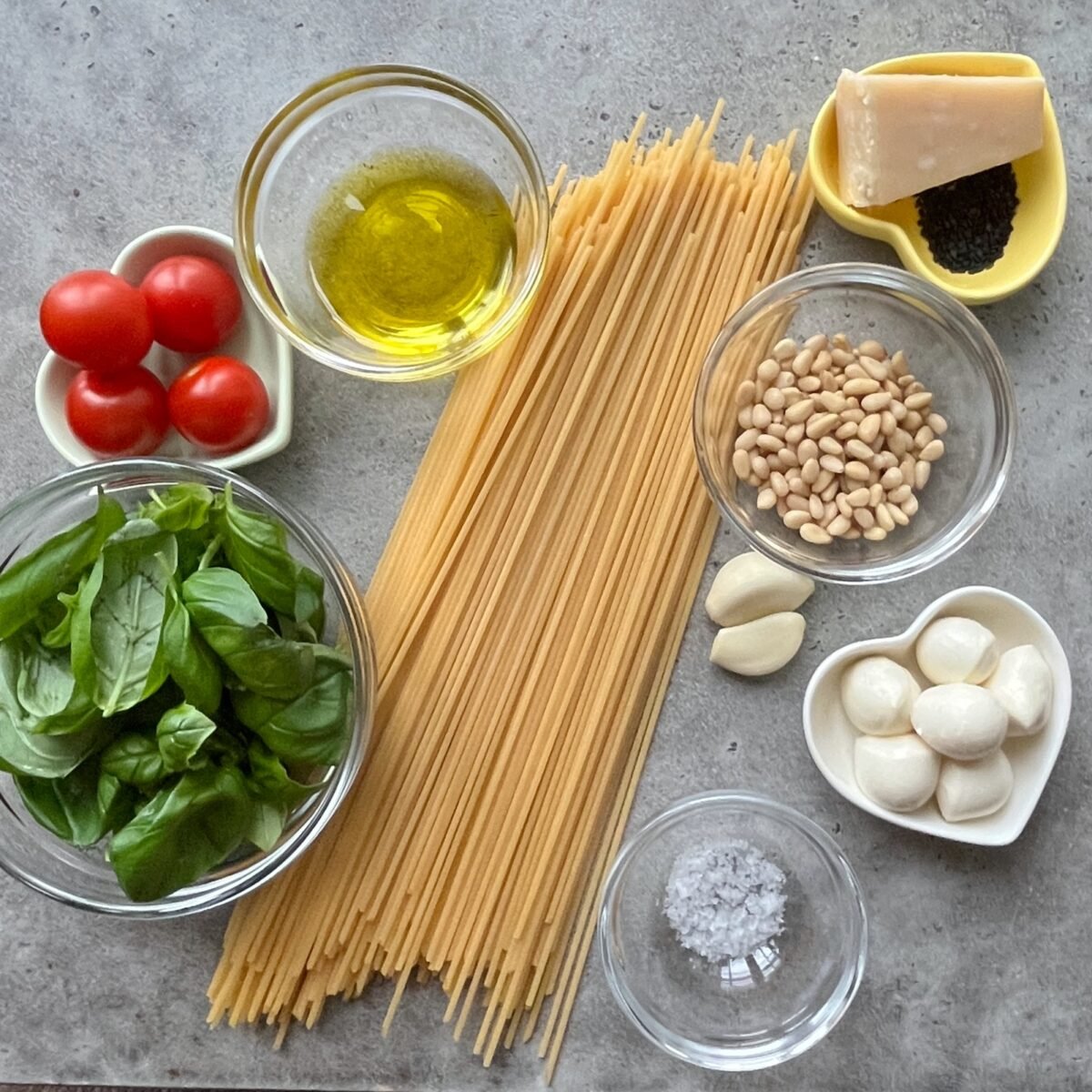 Various ingredients for an Italian pasta dish, including uncooked spaghetti, cherry tomatoes, basil, olive oil, pine nuts, garlic, mozzarella balls, Parmesan, salt, and black sesame seeds.