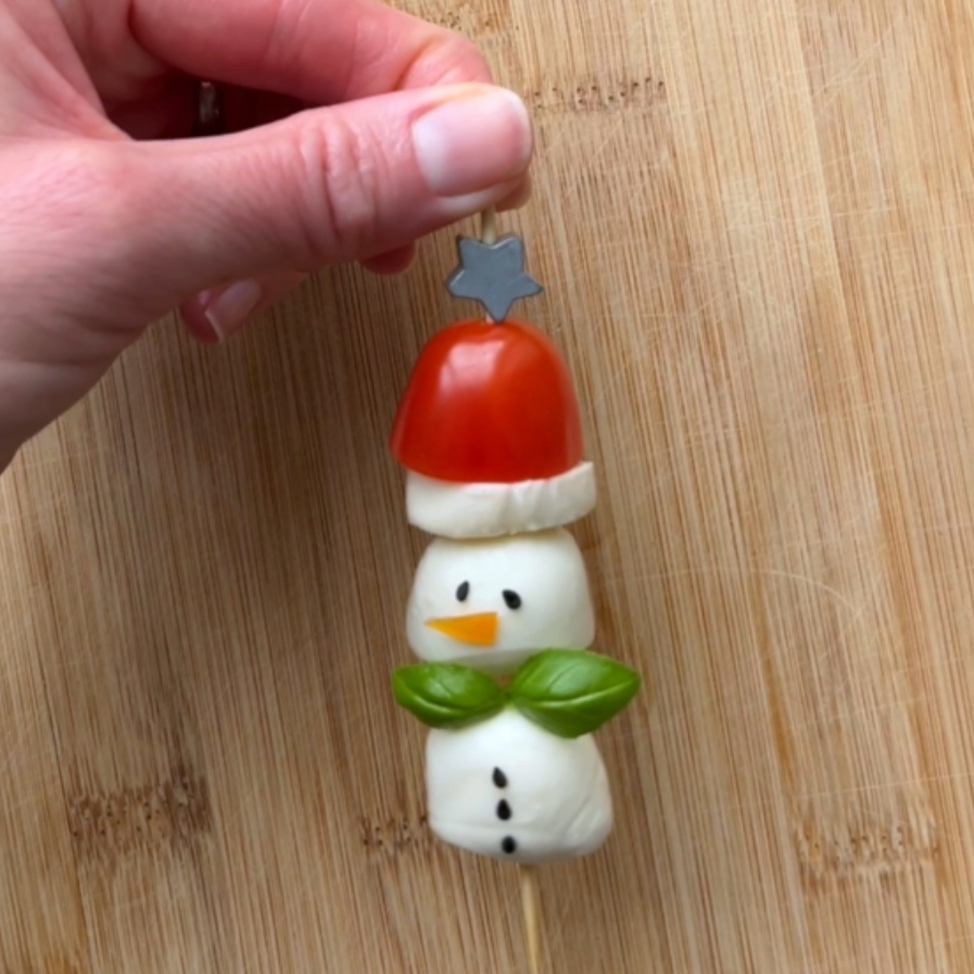 A hand holds a skewer with a snowman made of mozzarella balls, a tomato, basil leaves, and a carrot piece on a wooden surface.