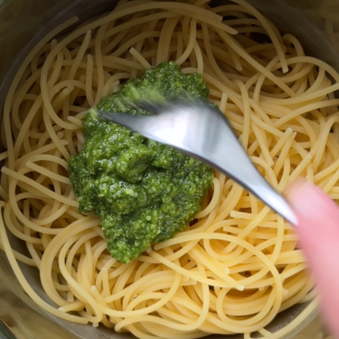 A hand spoons green basil pesto onto spaghetti in a metal bowl.