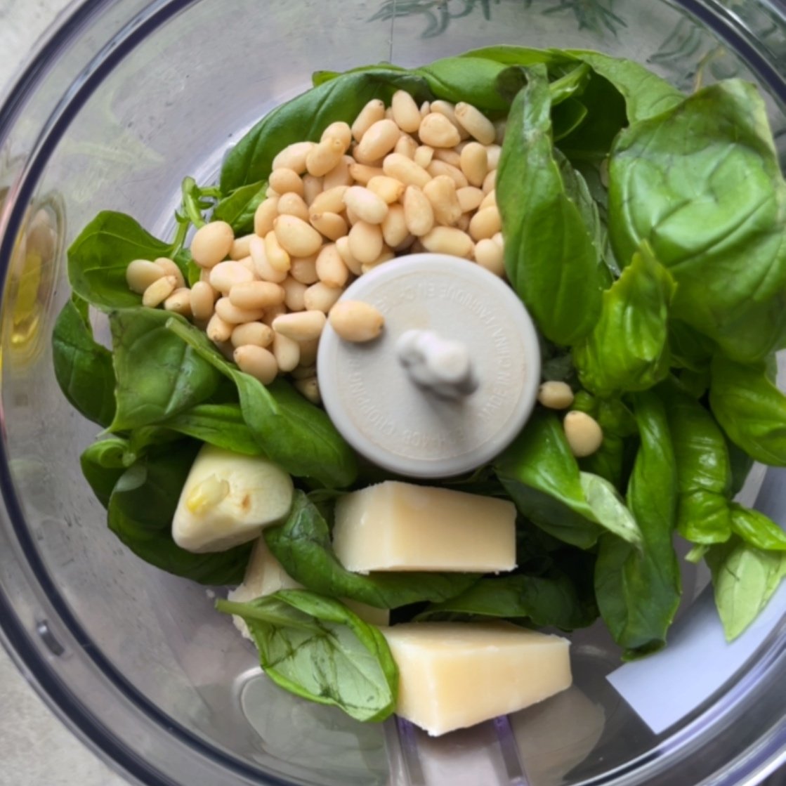 Basil leaves, pine nuts, garlic cloves, and Parmesan cheese in a food processor ready for blending.
