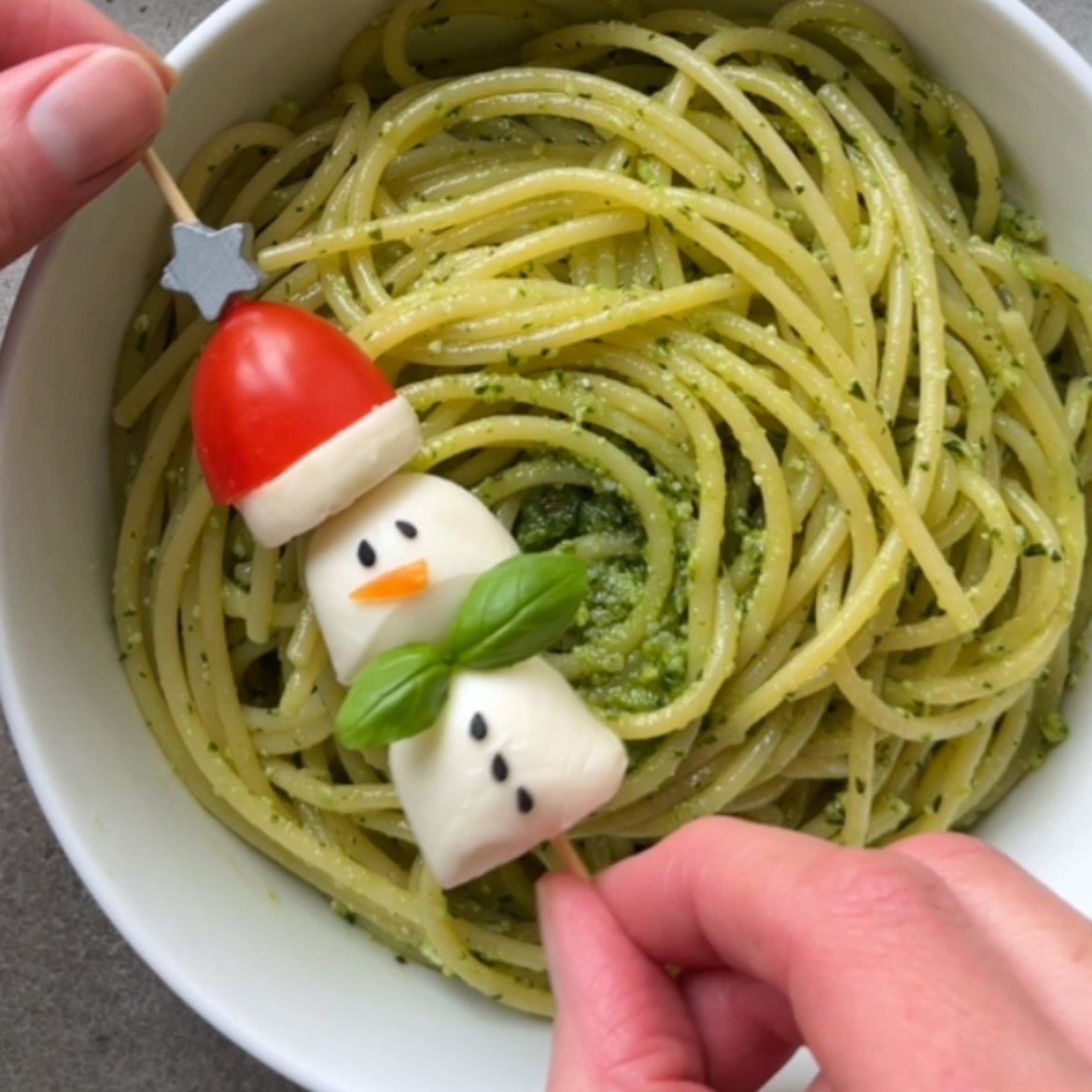 A festive bowl of pesto spaghetti topped with a skewer featuring a snowman made of mozzarella, tomato, and basil.
