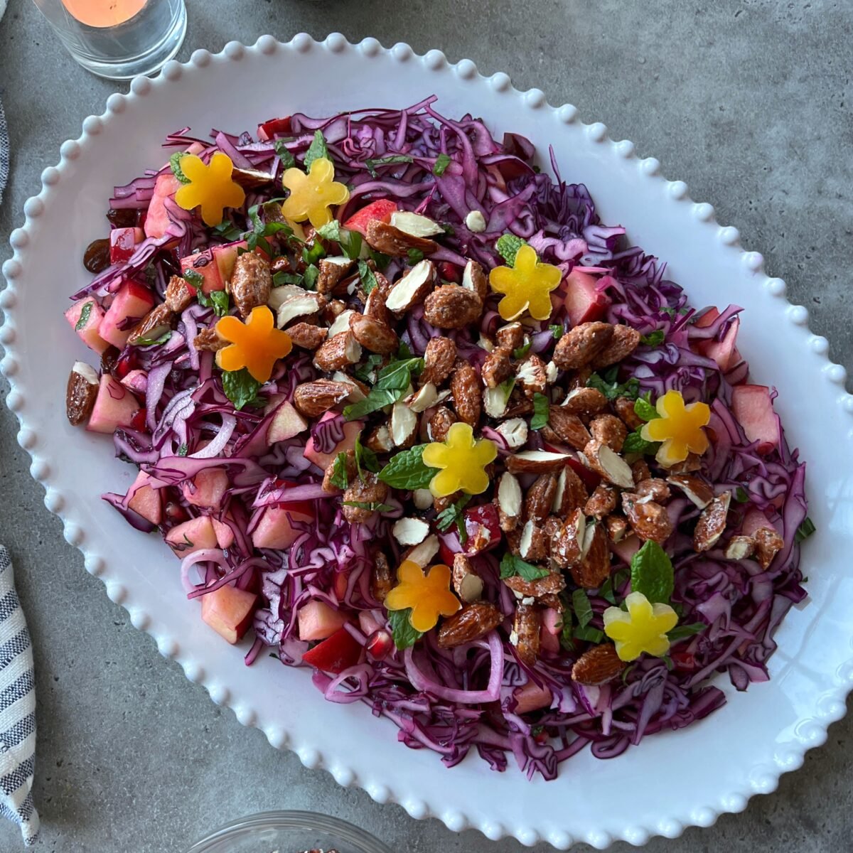 A side view of a salad on a white oval plate with shredded red cabbage, chopped apples, candied nuts, herbs, and yellow flower-shaped pieces, garnished on a gray surface.
