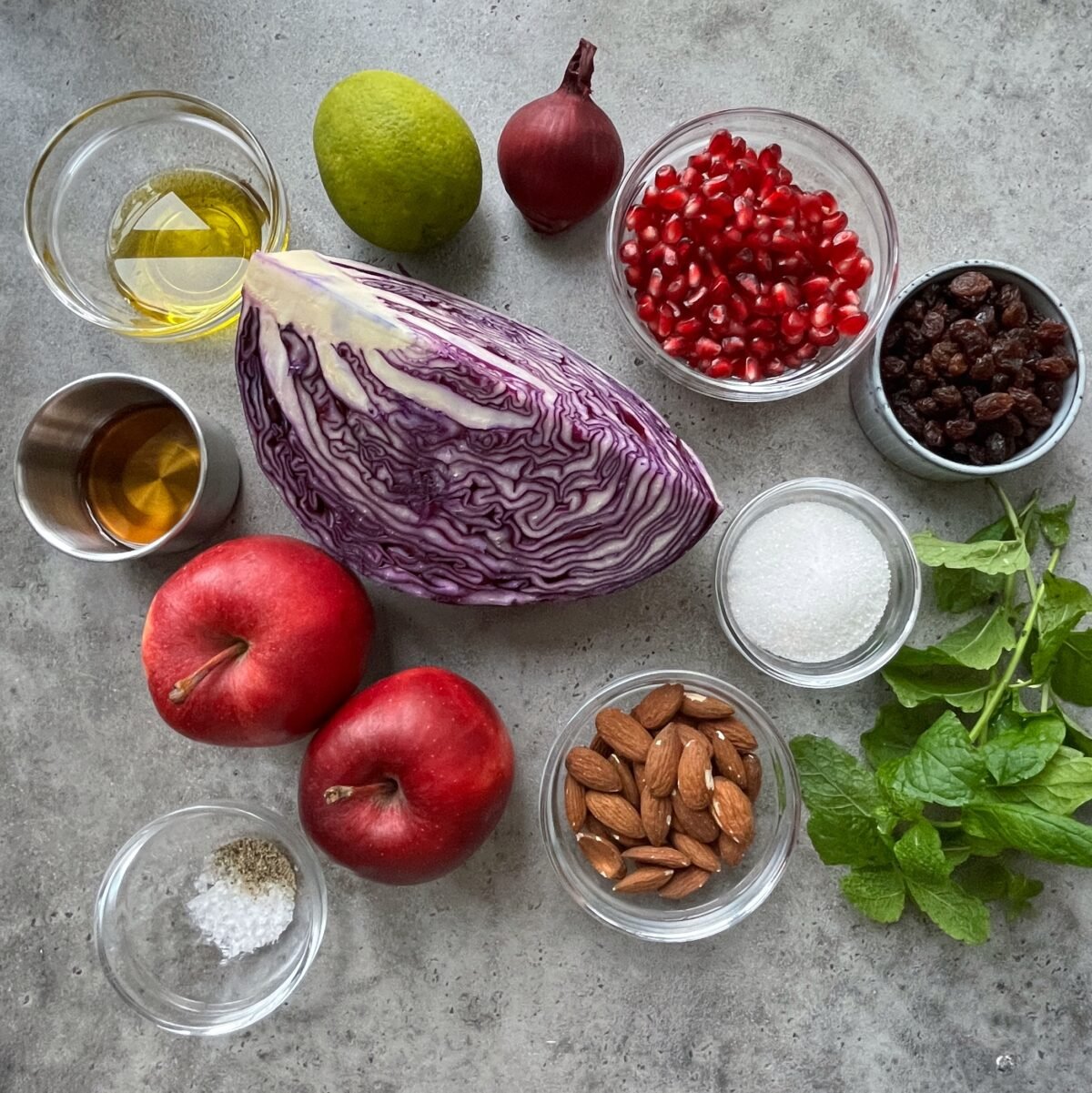 A flat lay of ingredients on a gray surface, including red cabbage, apples, pomegranate seeds, raisins, almonds, a red onion, mint, lime, oil, maple syrup, sugar, salt, and pepper.