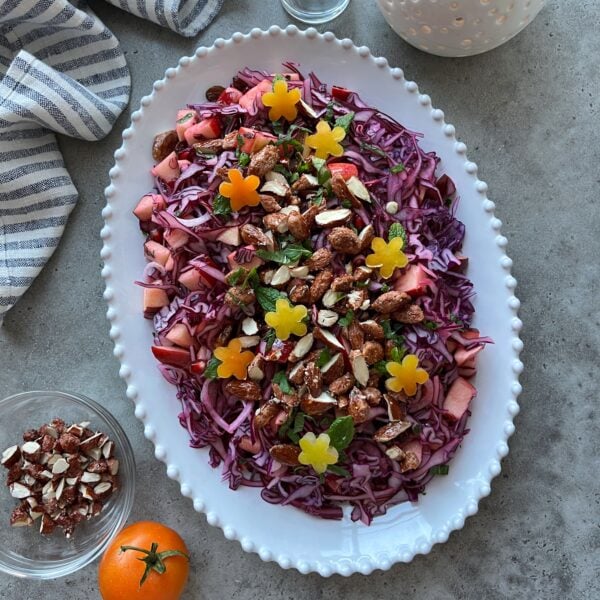A white oval platter with shredded red cabbage salad topped with chopped nuts, apple pieces, herbs, and yellow flower-shaped tomatoes on a gray surface.