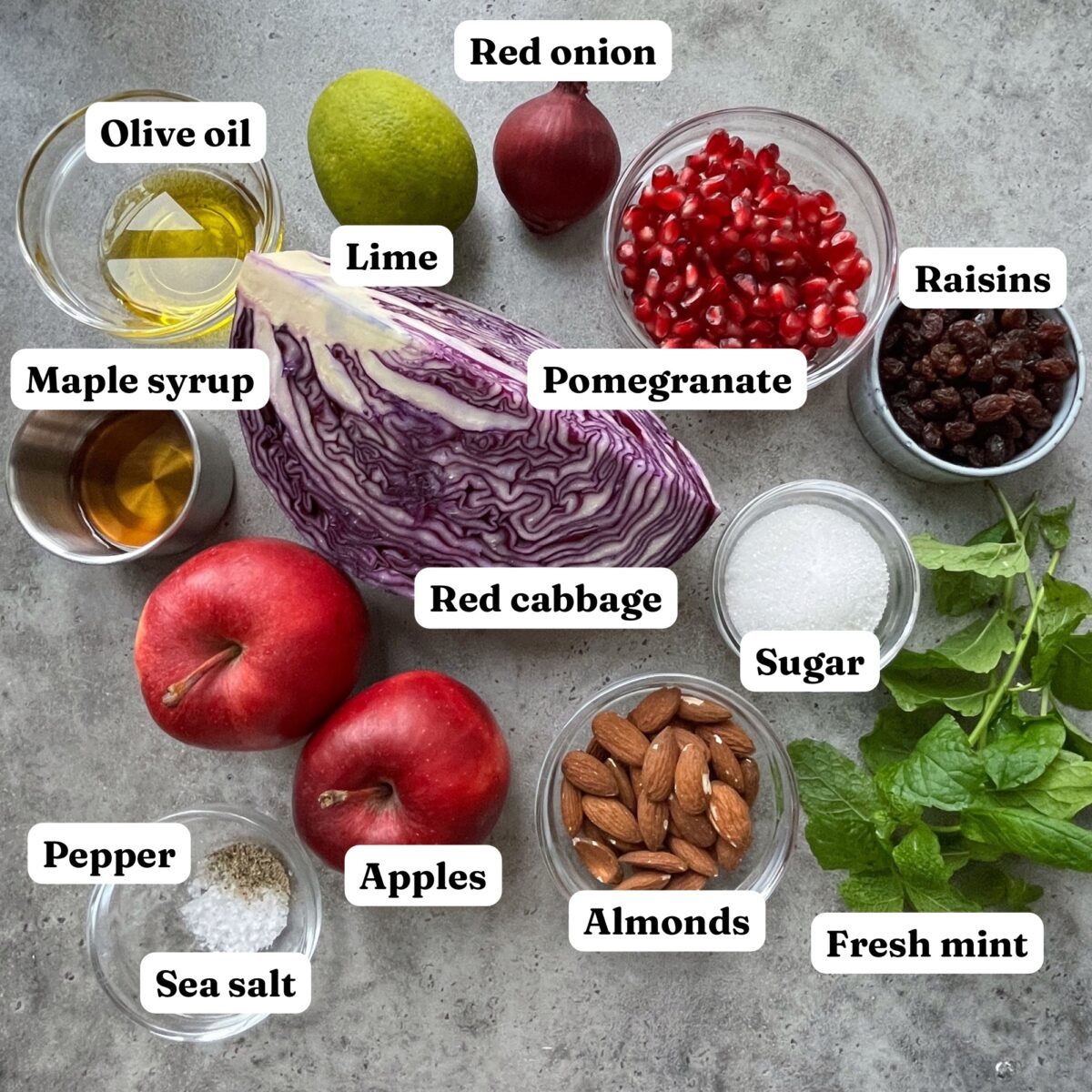 Assorted ingredients for a festive Christmas Salad are arranged on a gray surface: sliced red cabbage, apples, lime, red onion, olive oil, maple syrup, pomegranate, raisins, sugar, sea salt, pepper, almonds, and fresh mint.