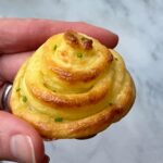 A hand holding a small, swirled Duchess Potatoes dish topped with chopped chives against a neutral background.