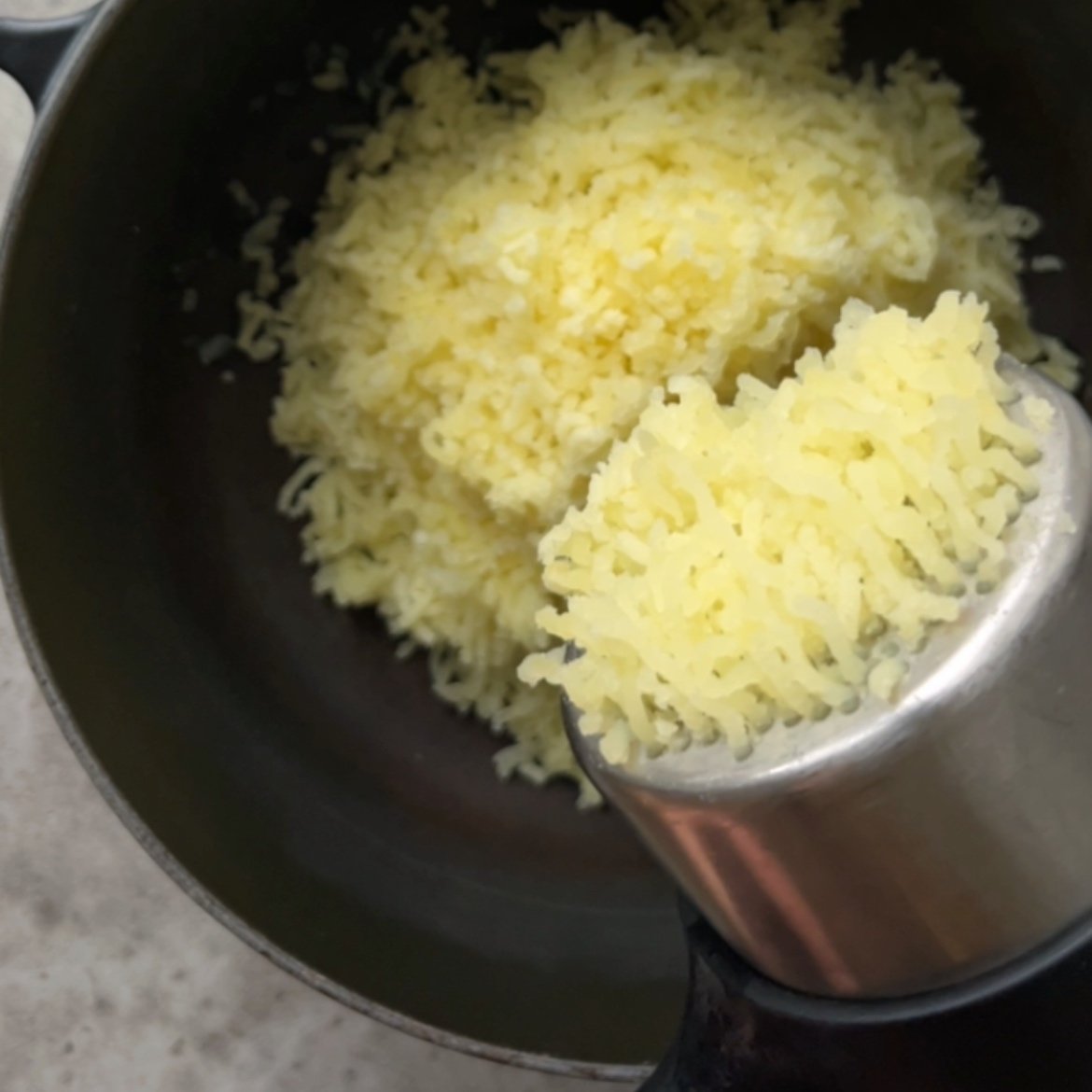 Mashed potatoes being pressed through a potato ricer into a black bowl, with some potato still in the ricer.