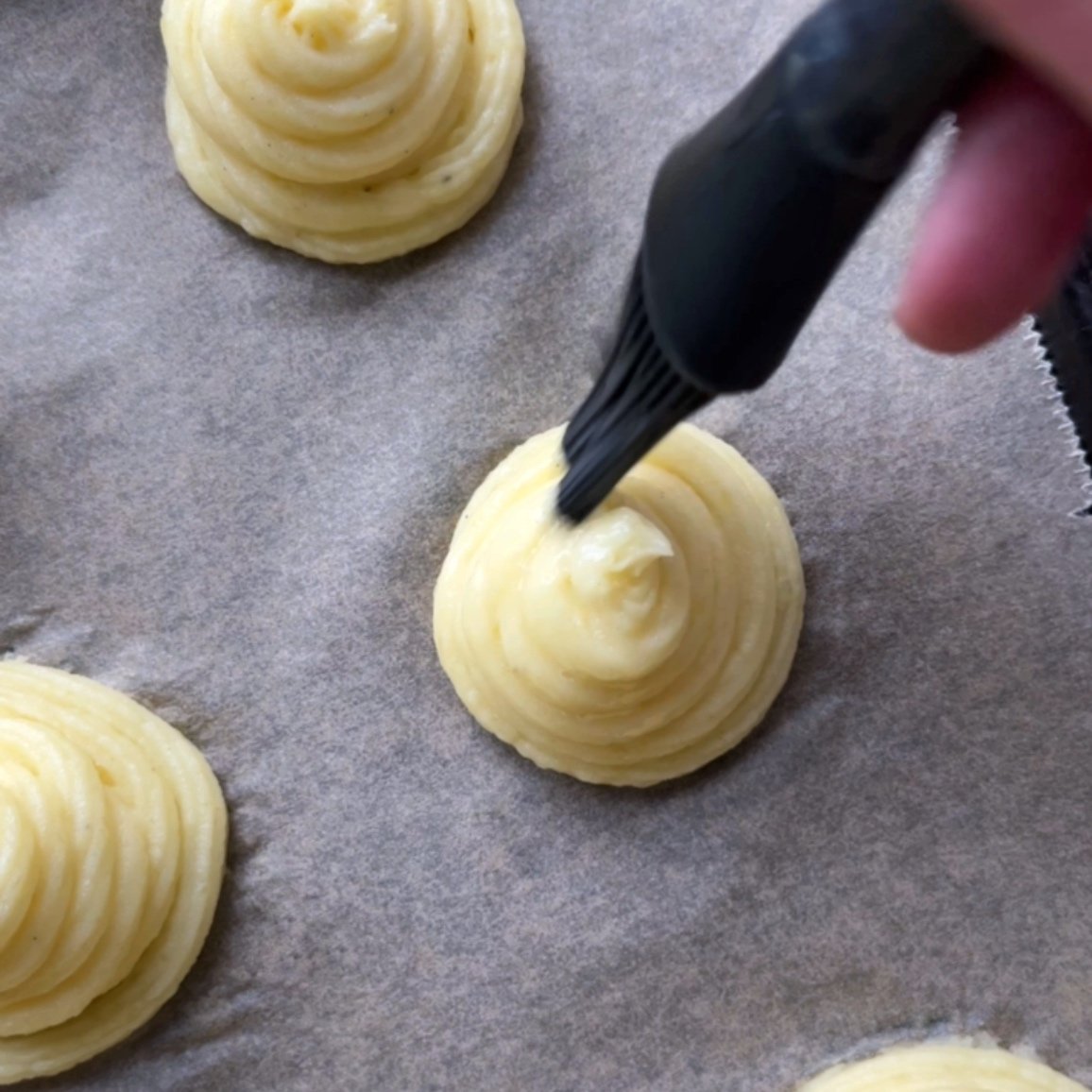 A hand uses a pastry brush to apply egg wash to piped swirls of Duchess Potatoes on parchment paper.