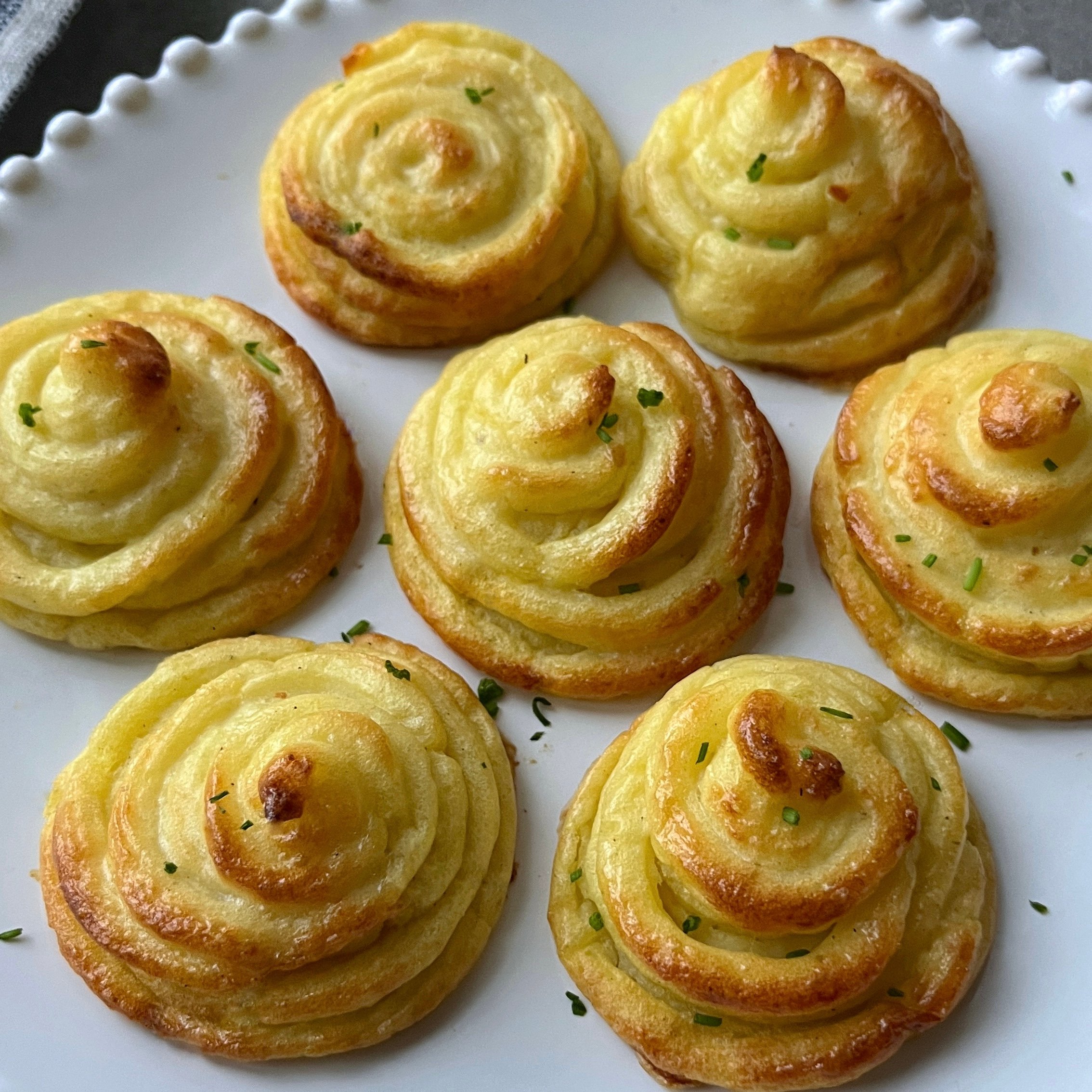 Seven piped duchess potatoes, golden brown and garnished with herbs, are arranged on a white plate with a scalloped edge.