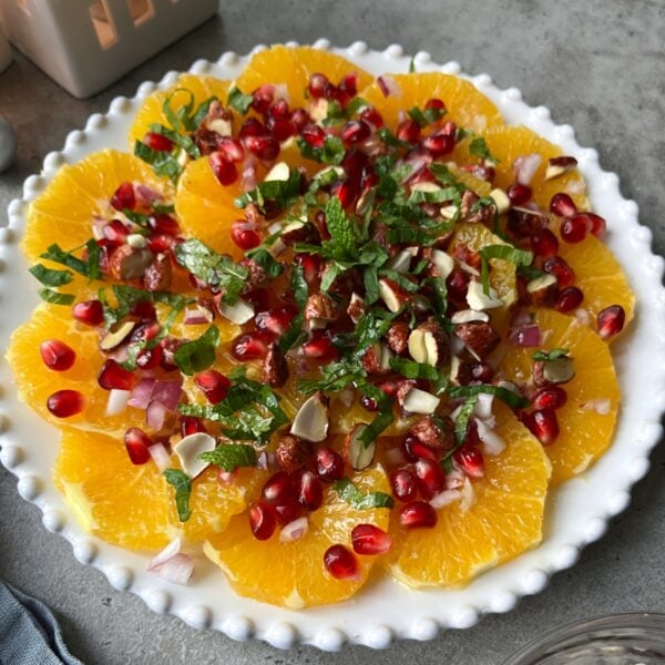 A plate of sliced oranges topped with pomegranate seeds, chopped nuts, diced red onions, and fresh mint, arranged on a white dish.