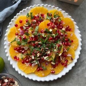 A plate of sliced oranges topped with pomegranate seeds, chopped nuts, fresh mint, and red onion on a gray surface.
