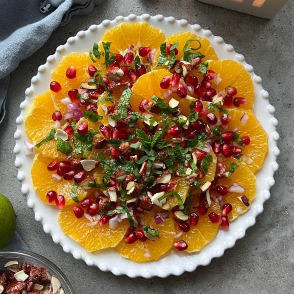 A plate of sliced oranges topped with pomegranate seeds, chopped nuts, fresh mint, and red onion on a gray surface.