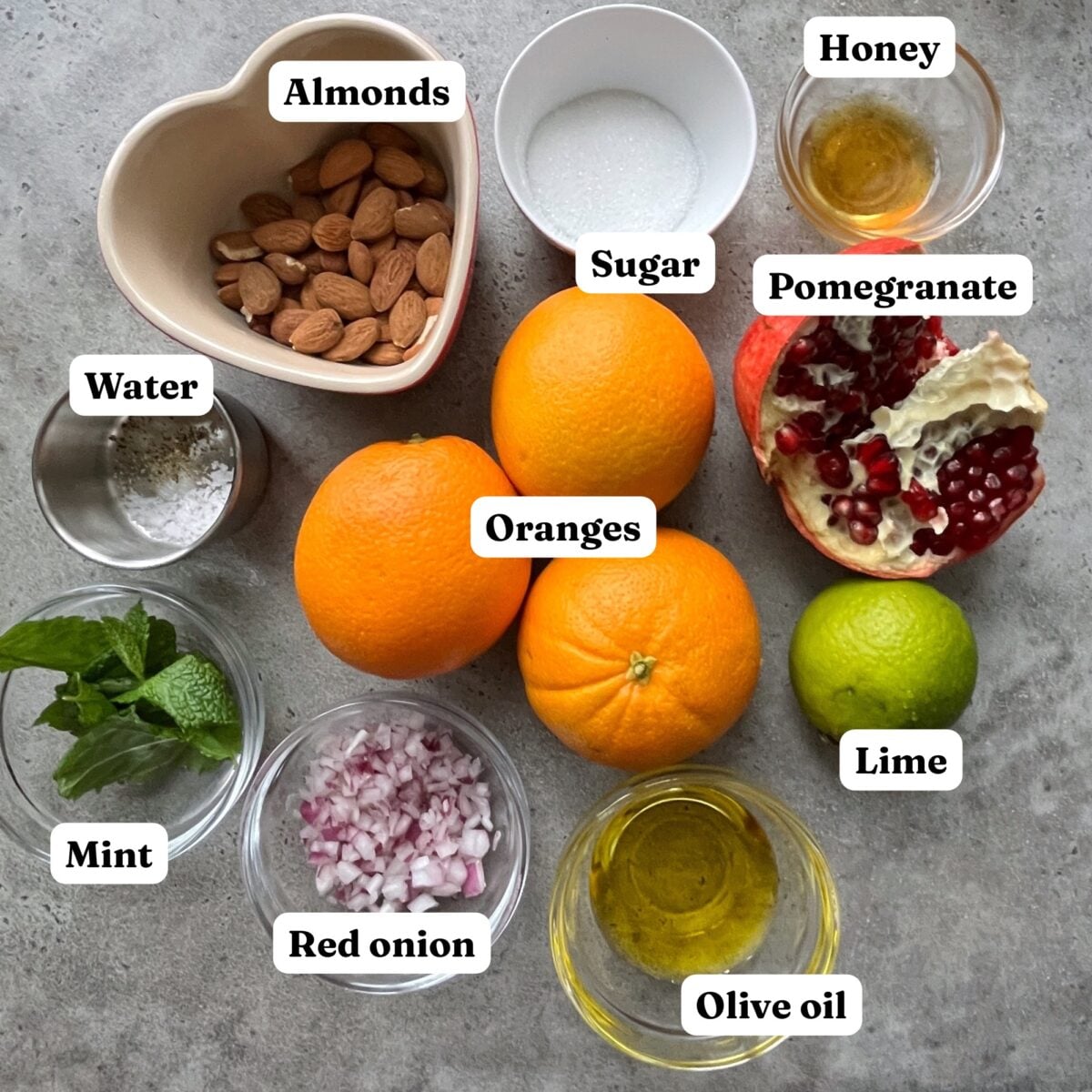 A flat lay of ingredients in bowls, including almonds, honey, sugar, pomegranate, water, oranges, lime, mint, red onion, and olive oil.
