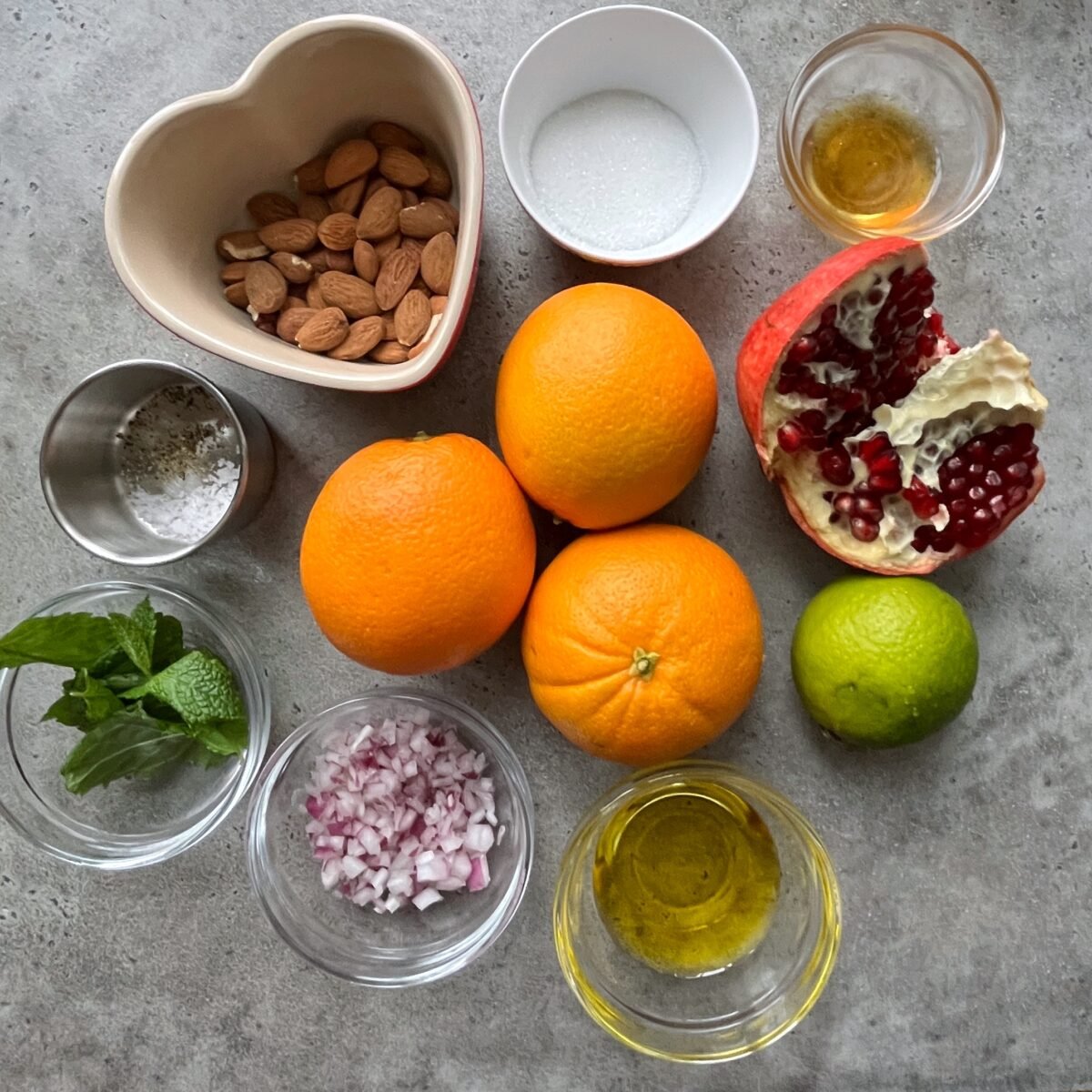 A flat lay of almonds, sugar, oranges, pomegranate, lime, fresh mint, chopped red onion, olive oil, salt, and honey in small bowls on a gray surface.