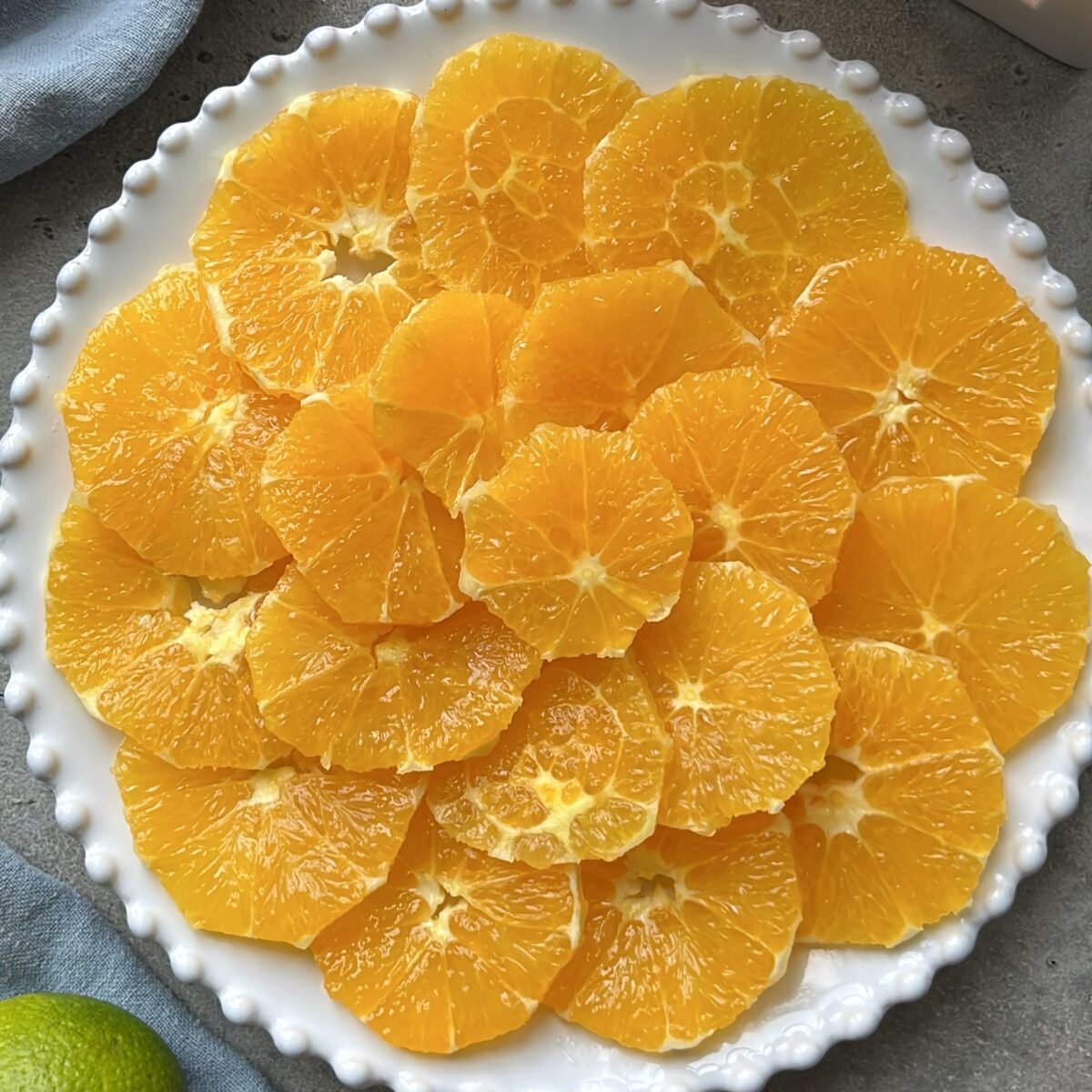 A white plate with a raised beaded edge holding neatly arranged slices of peeled orange fruit.