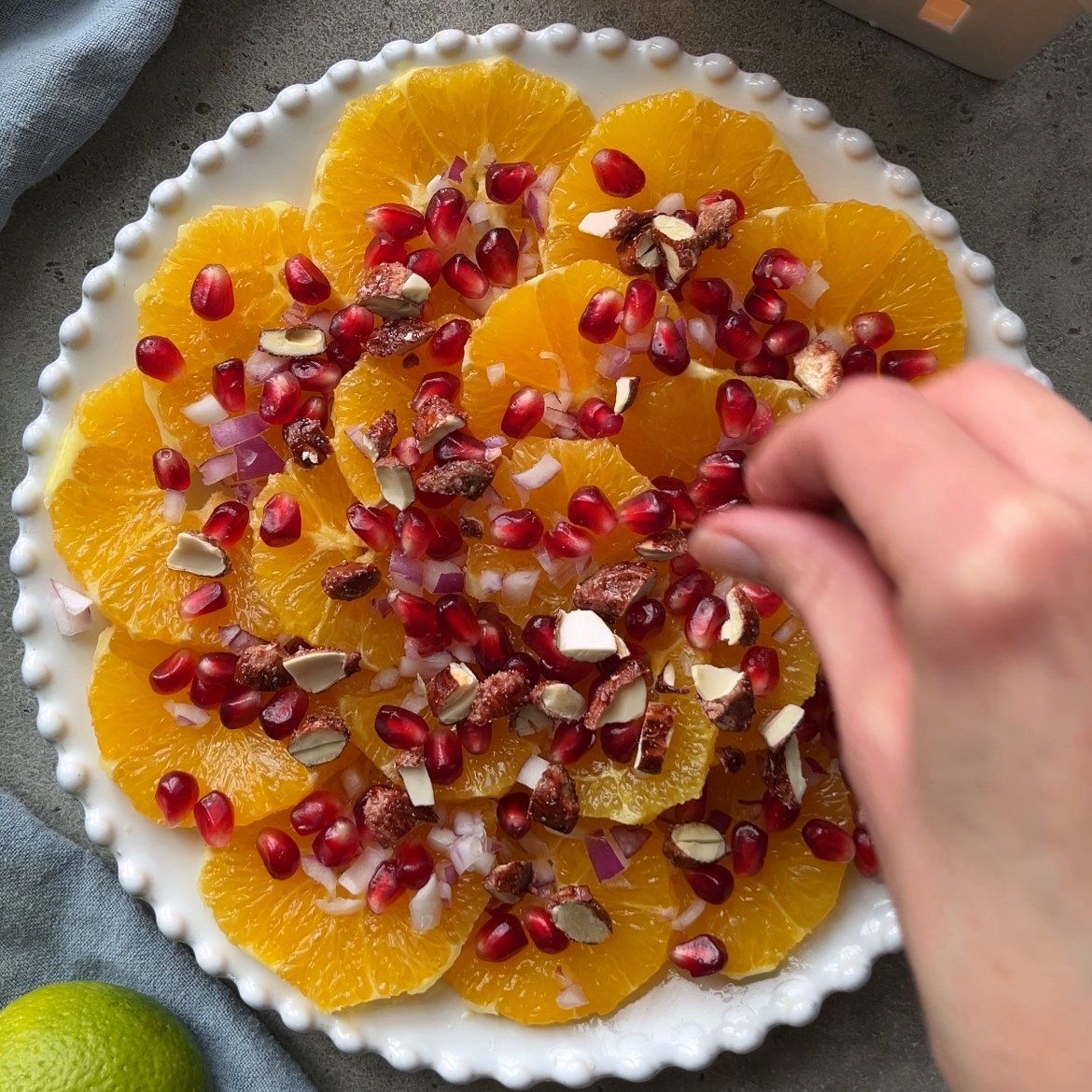 A hand sprinkles toppings over a plate of sliced oranges garnished with pomegranate seeds, chopped nuts, and diced red onions.