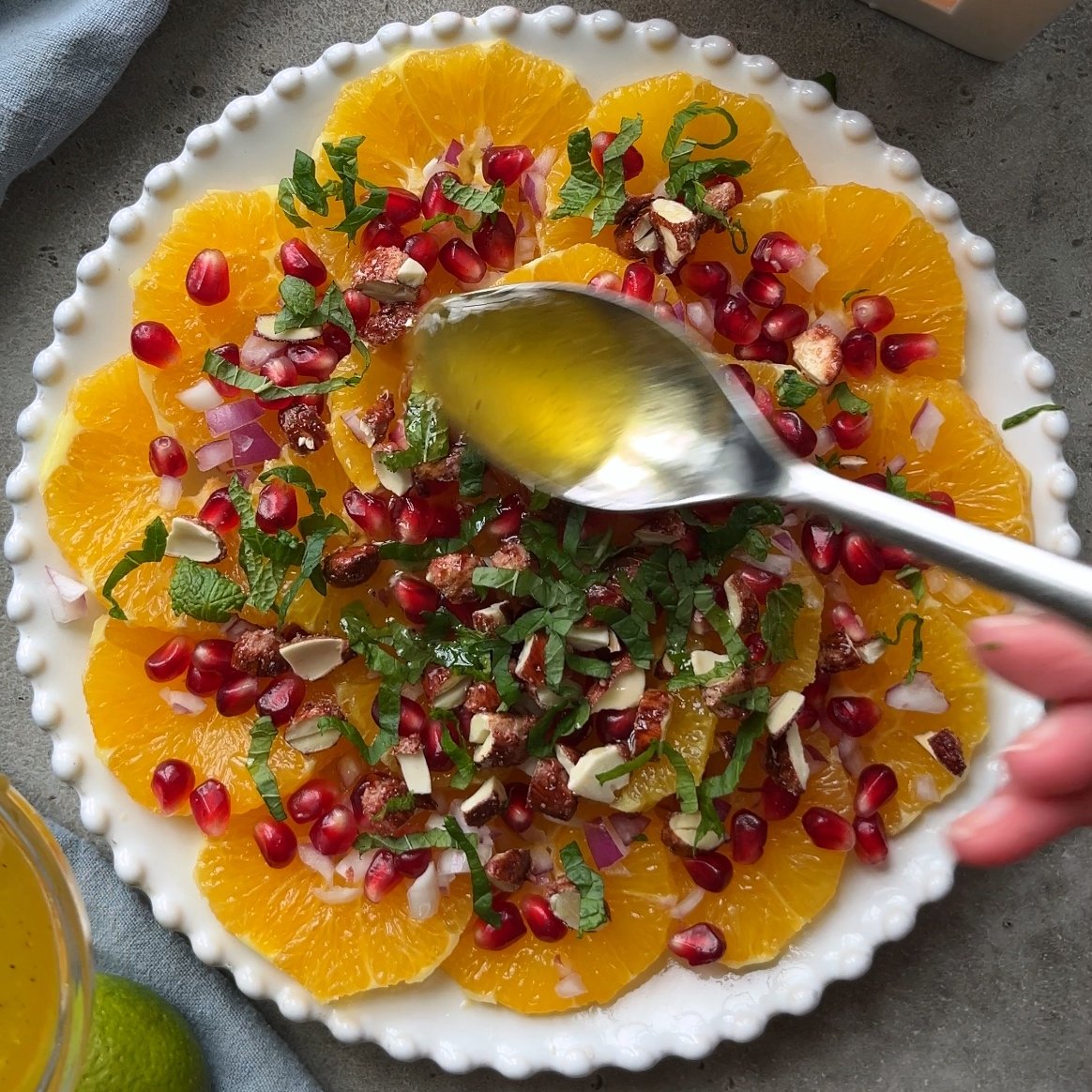 A hand drizzles dressing over a plate of sliced oranges topped with pomegranate seeds, chopped nuts, mint, and diced red onion.