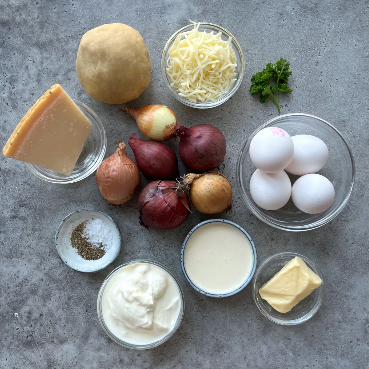 Various ingredients on a gray surface, including eggs, onions, cheeses, cream, butter, pastry dough for Caramelized Onion Quiche, yogurt, salt, pepper, and a sprig of parsley.