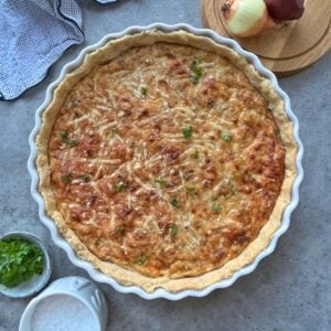 A baked Caramelized Onion Quiche with a golden, cheesy top sits in a white pie dish, garnished with herbs. An onion, parsley, and a bowl of salt are nearby on a gray surface.