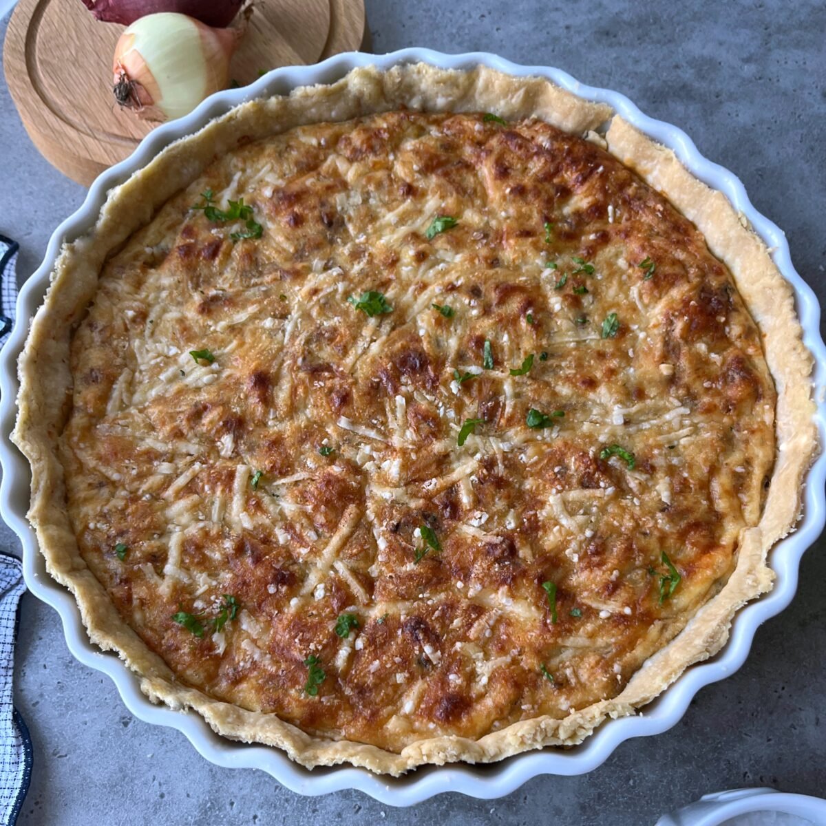 A baked Caramelised Onion Quiche with a golden, cheesy top is shown in a round dish, garnished with herbs. An onion and a cutting board are visible in the background.