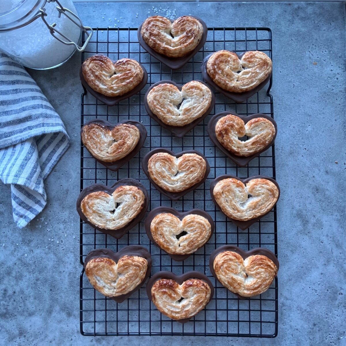 A cooling rack holds thirteen heart-shaped pastries on a gray countertop, with a striped towel and a jar of sugar nearby.