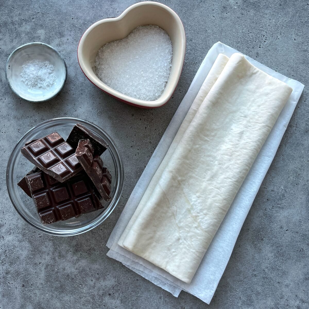 A bowl of chocolate pieces, a folded sheet of puff pastry, a heart-shaped bowl of coarse sugar, and a small bowl of salt on a gray surface.