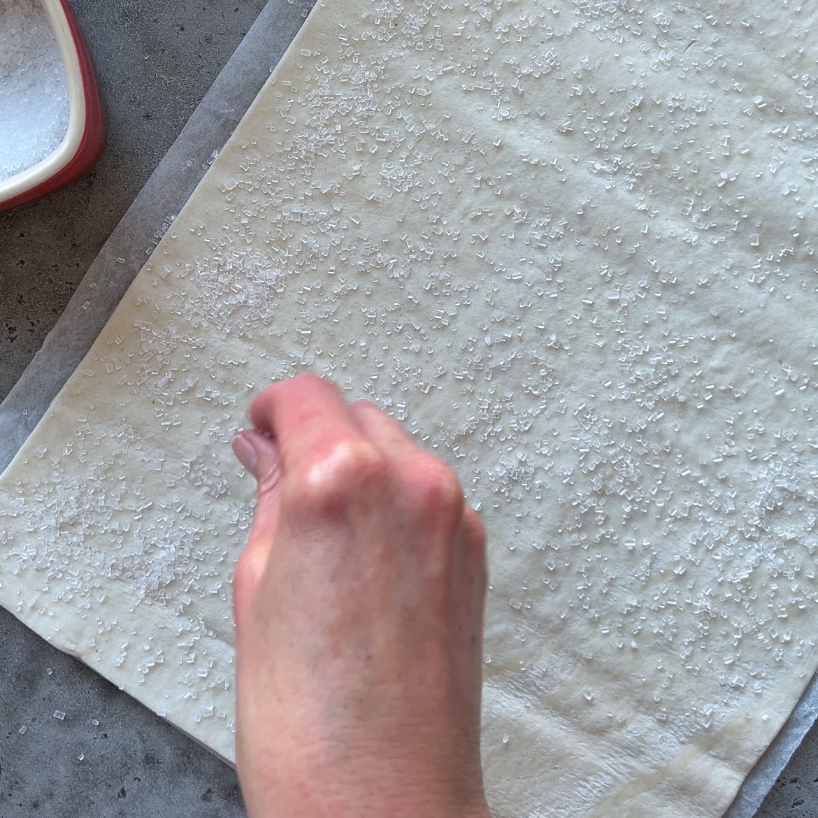 A hand sprinkles coarse sugar onto a sheet of pastry dough placed on parchment paper.