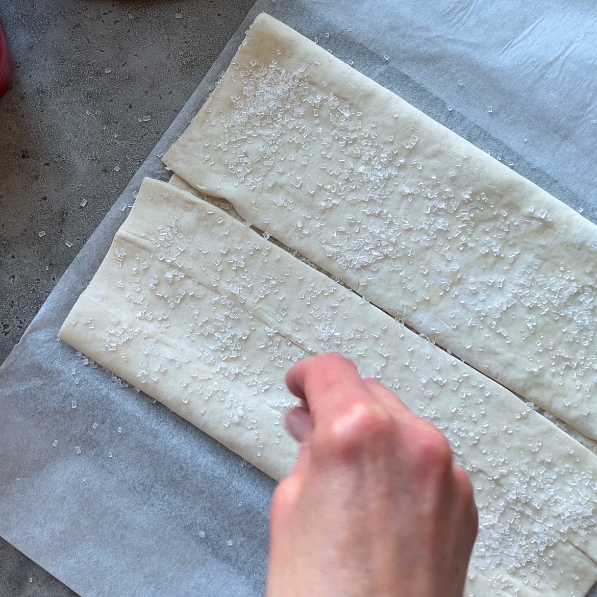 A hand sprinkles coarse sugar onto folded puff pastry dough placed on parchment paper.