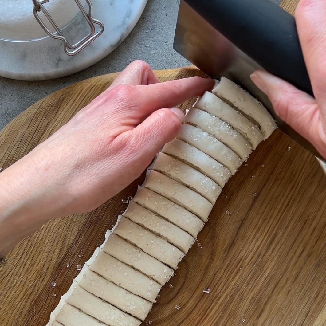 A person uses a bench scraper to cut a rolled dough log into even slices on a wooden board.