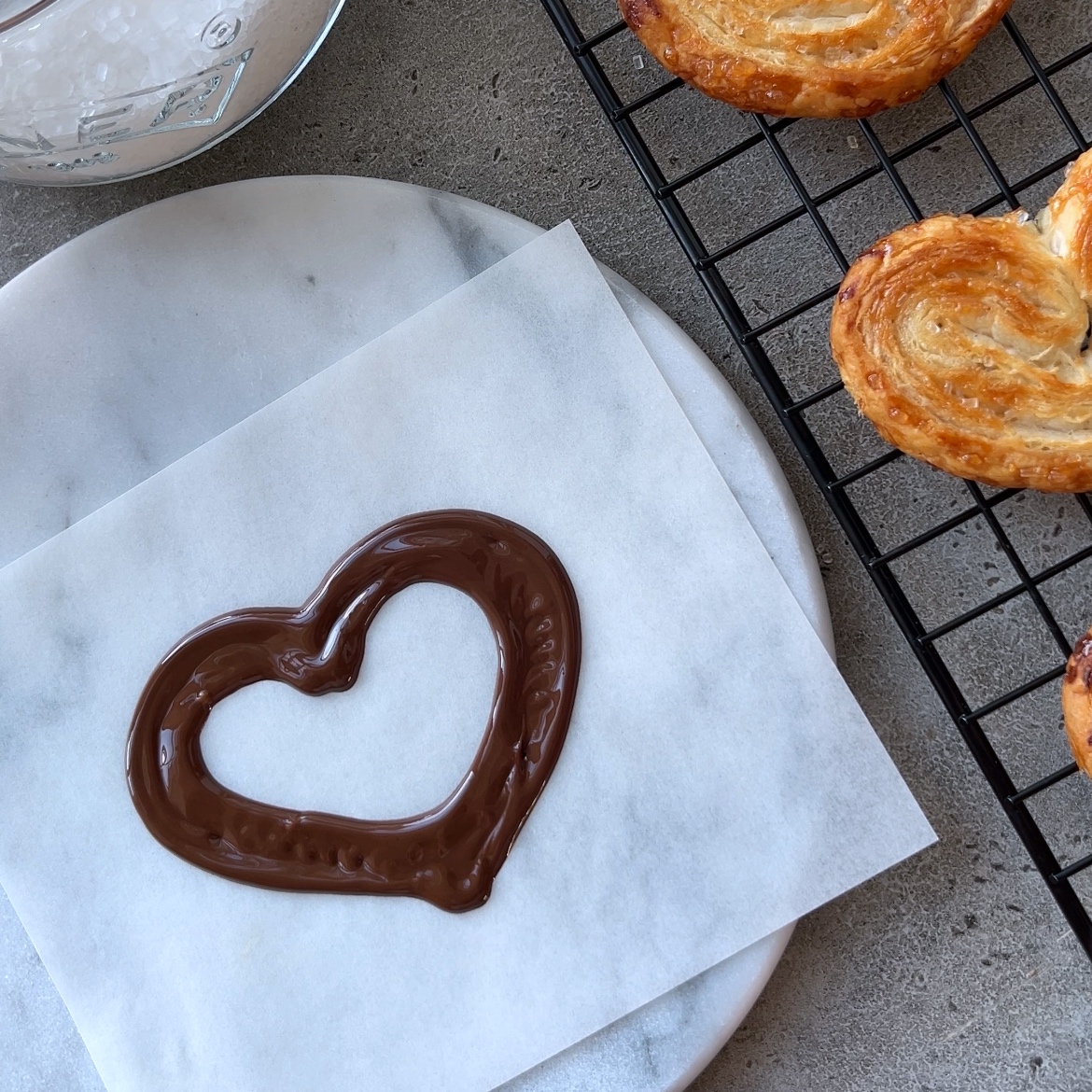 A heart shape drawn with chocolate on parchment paper, placed on a marble board next to a cooling rack with baked pastries.