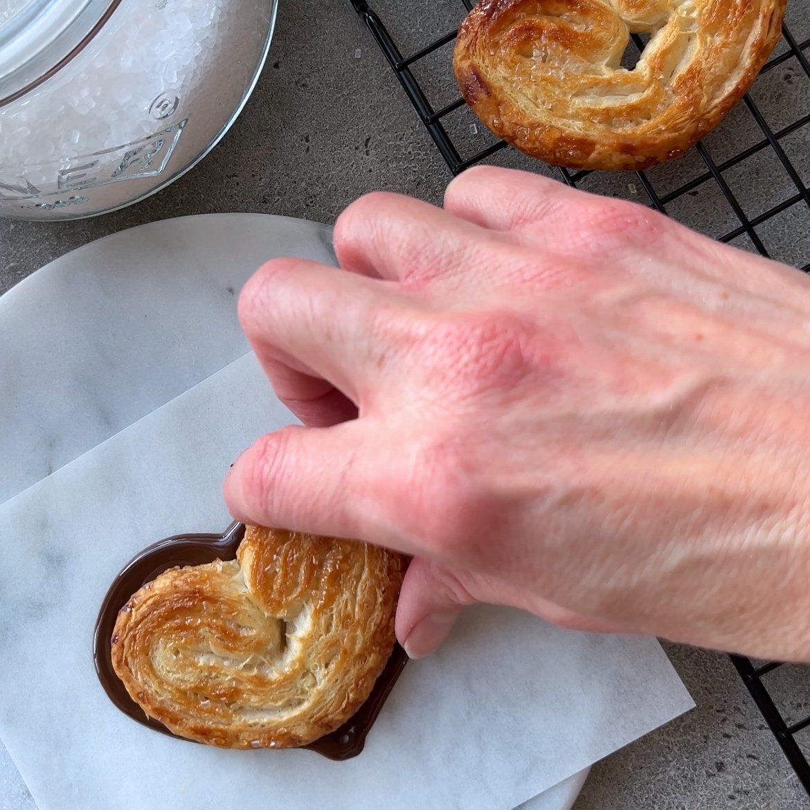 A hand places a heart-shaped puff pastry onto melted chocolate on parchment paper.