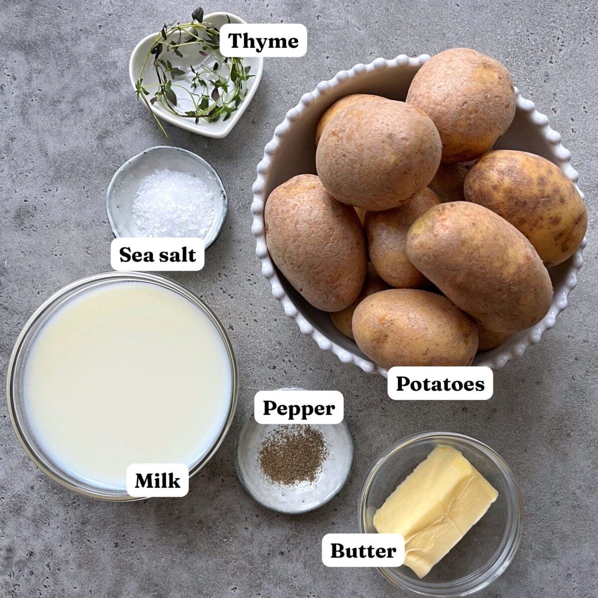 A bowl of potatoes, a dish of butter, a cup of milk, a small bowl of sea salt, pepper, and sprigs of thyme are arranged on a gray surface.