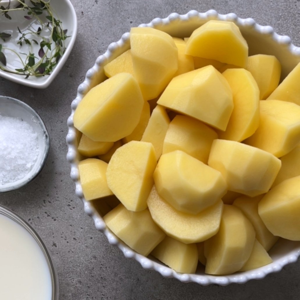 A bowl of peeled and chopped potatoes next to a dish of salt, a bowl of milk, and a small plate with fresh thyme on a gray countertop.