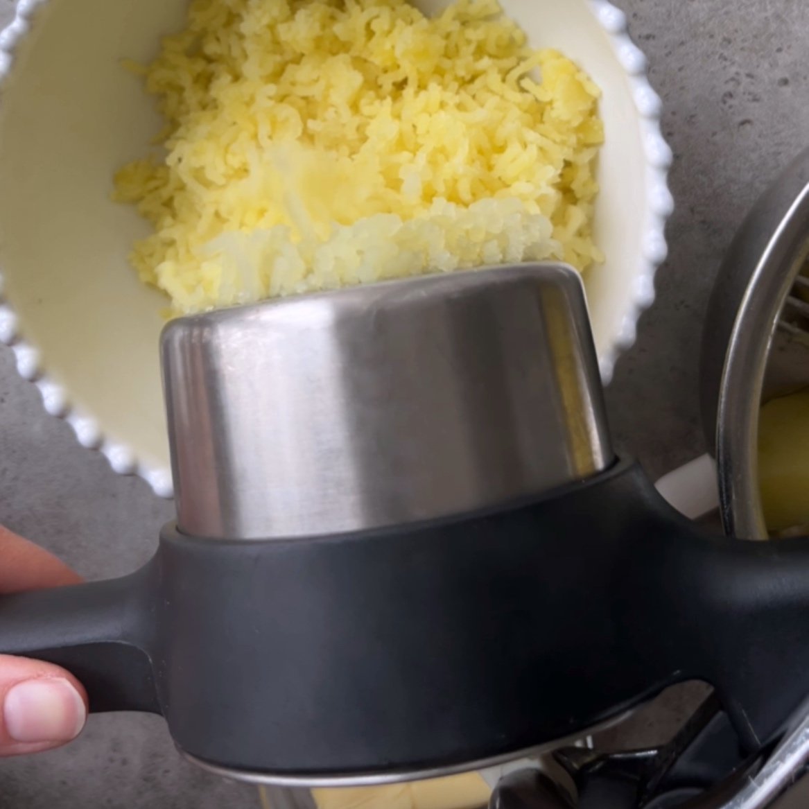 A hand holds a potato ricer over a bowl with riced potatoes, with more  potato chunks visible in a metal bowl.