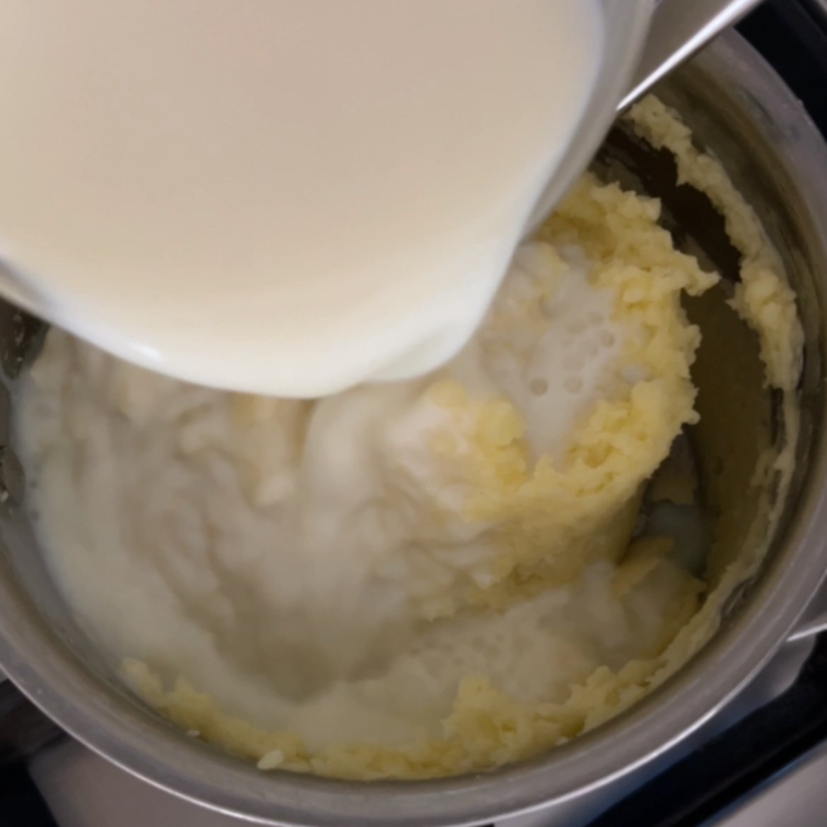 A metal pot with mashed potatoes inside as milk is being poured into them from a measuring cup.
