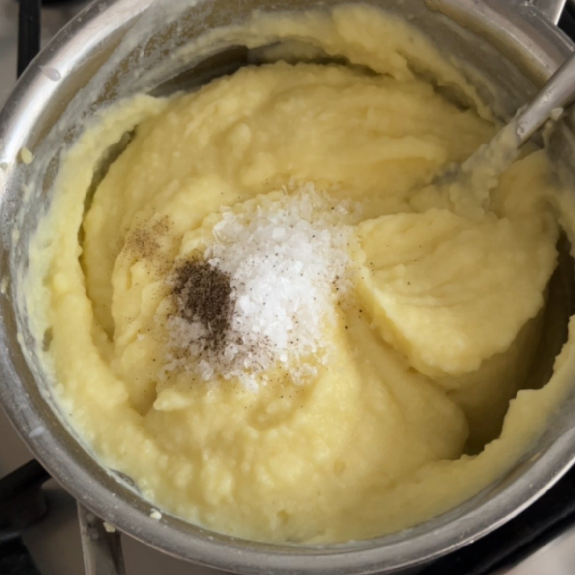 A pot of mashed potatoes topped with a small pile of salt and ground black pepper, and a metal spoon partially visible on the right.