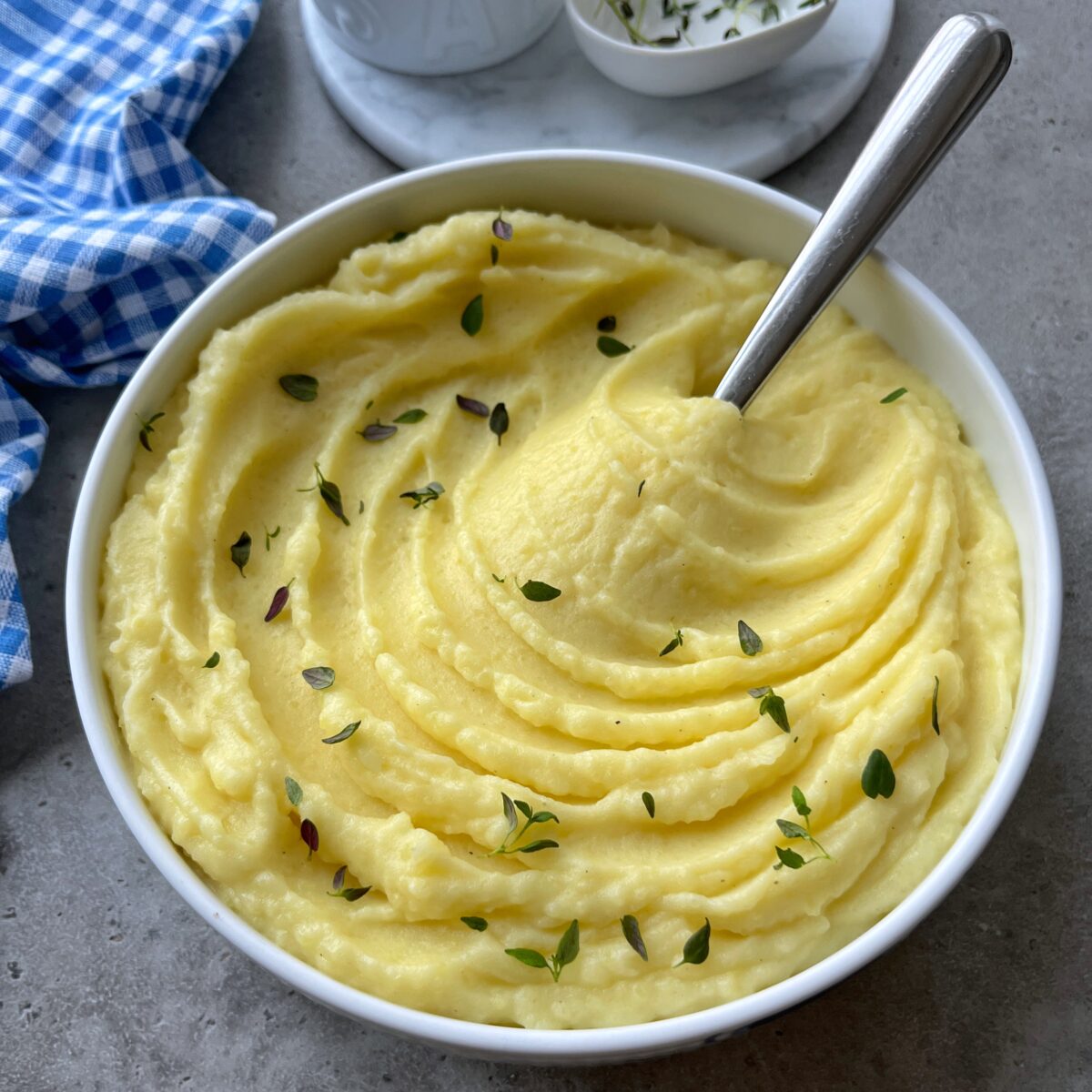 A side view of a bowl of mashed potatoes garnished with fresh herbs, with a spoon inside. 